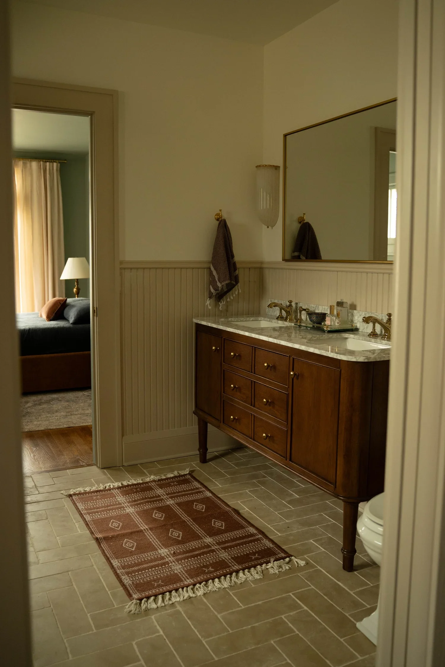 Bathroom with a wooden vanity, marble countertop, double sinks, large mirror, wall-mounted light fixture, towel hanging on a gold hook, patterned rug on beige tile floor, open door leading to a bedroom with curtains, bed, and lamp.