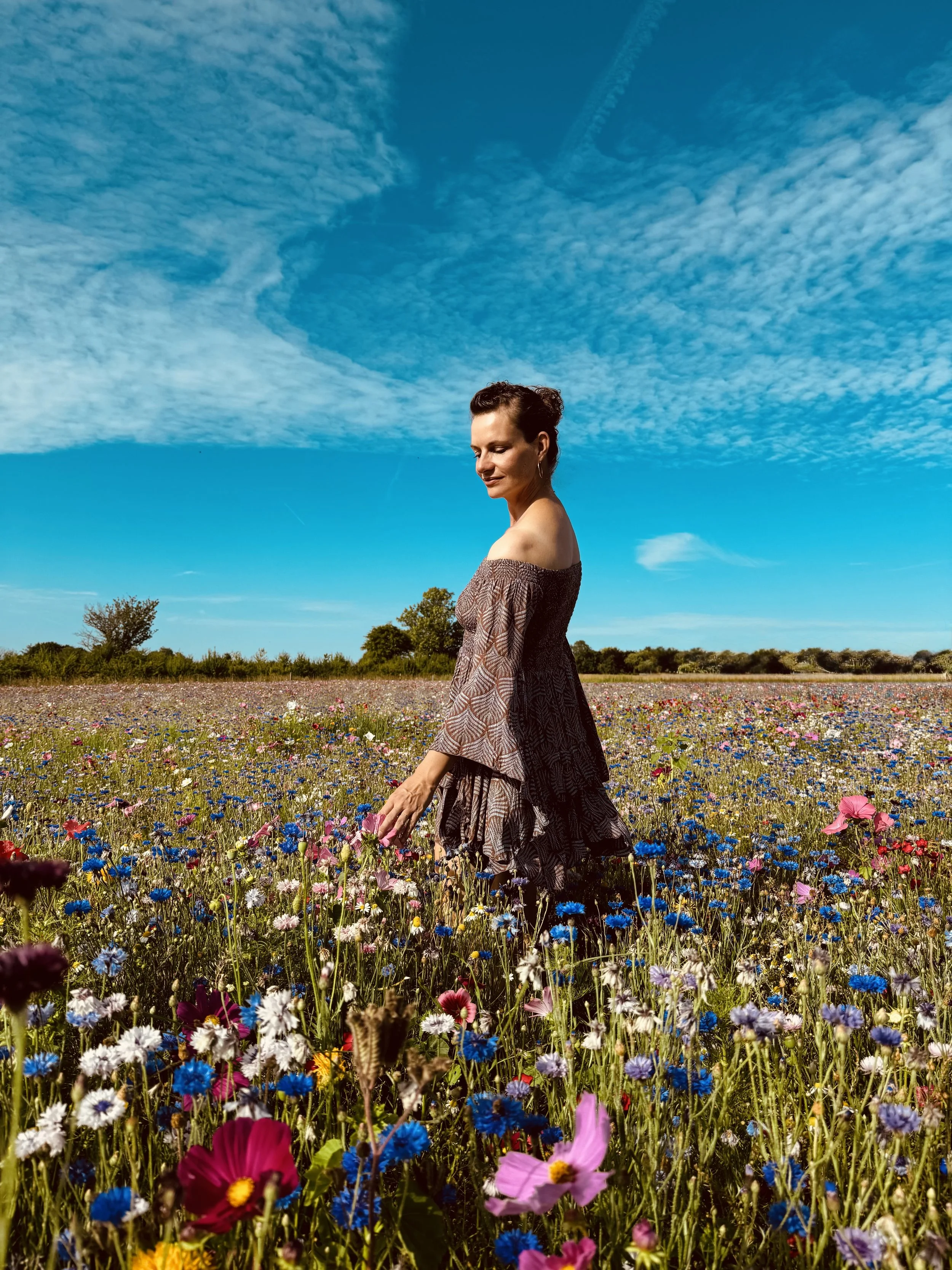 A woman in a patterned dress walking through a field of colorful wildflowers under a bright blue sky with scattered clouds.