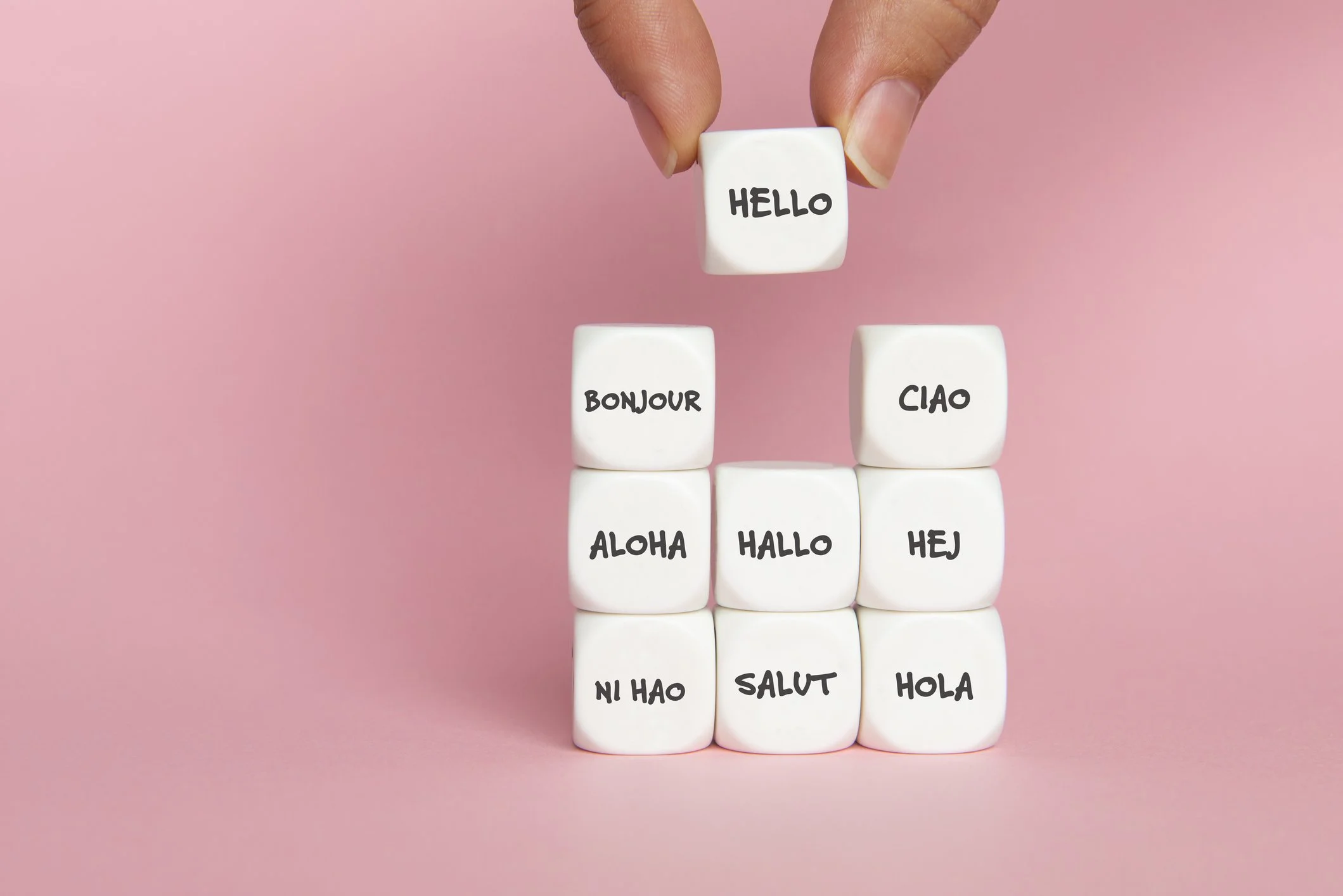 A hand stacking white dice with words for hello in different languages on a pink background.