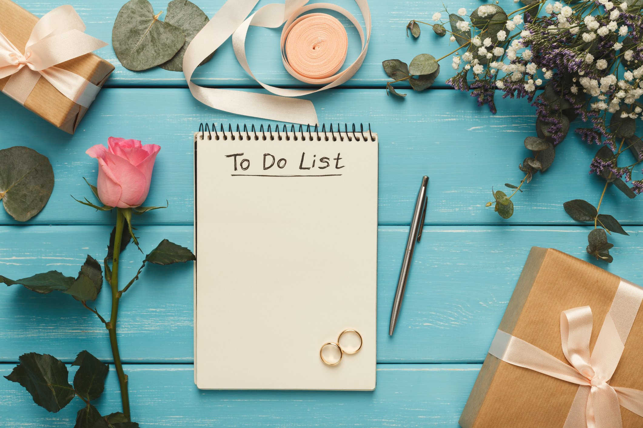 A to-do list notepad on a blue wooden table surrounded by pink rose, wrapped gifts, flowers, a pen, wedding rings, and decorative elements for a celebration or special occasion.