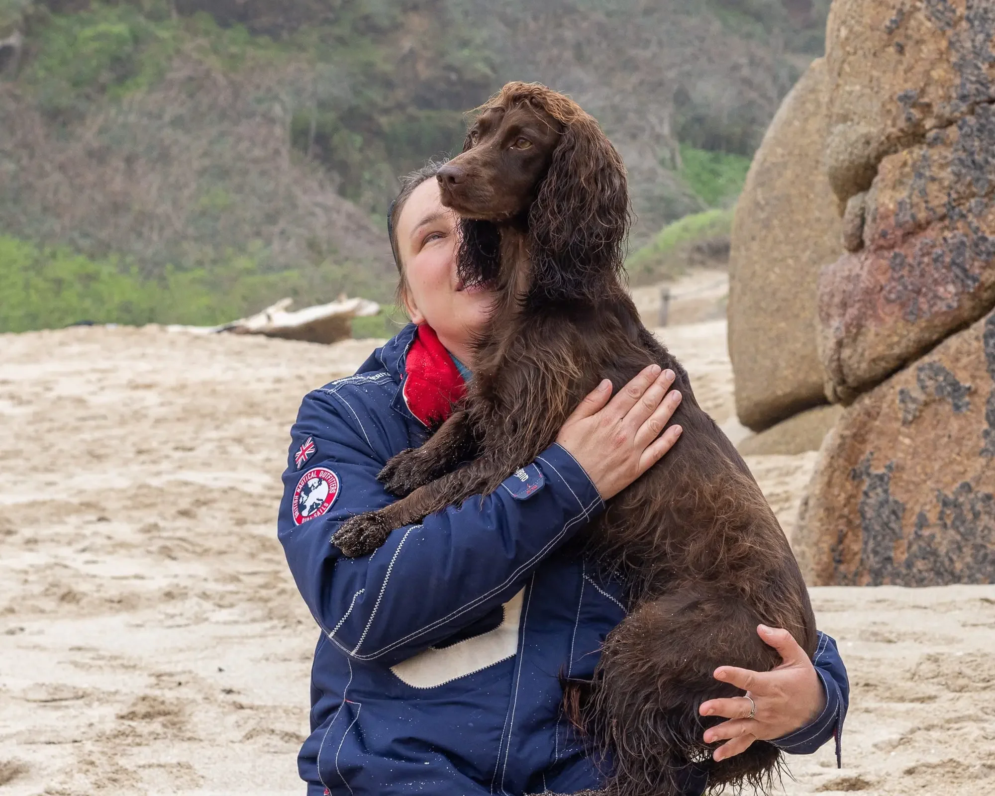 A woman hugging a brown spaniel on a beach, with rocks and green hills behind them, capturing the deep connection that guides her work with dogs.