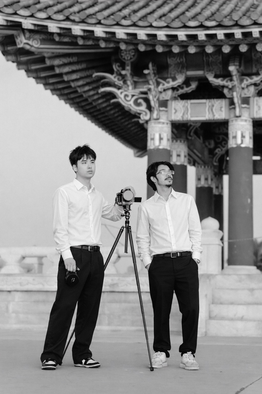 Two men in white shirts and black pants standing under an ornate, traditional Asian pagoda, one with a camera on a tripod and the other with his hands in his pockets, in black and white.