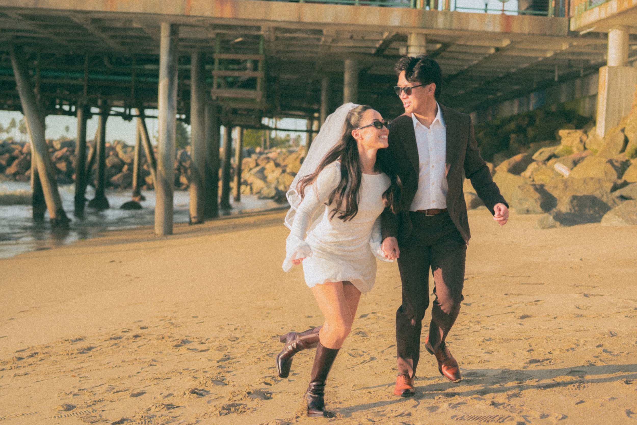 A couple dressed in wedding attire walking on the beach, smiling and holding hands, with a pier and rocks in the background.