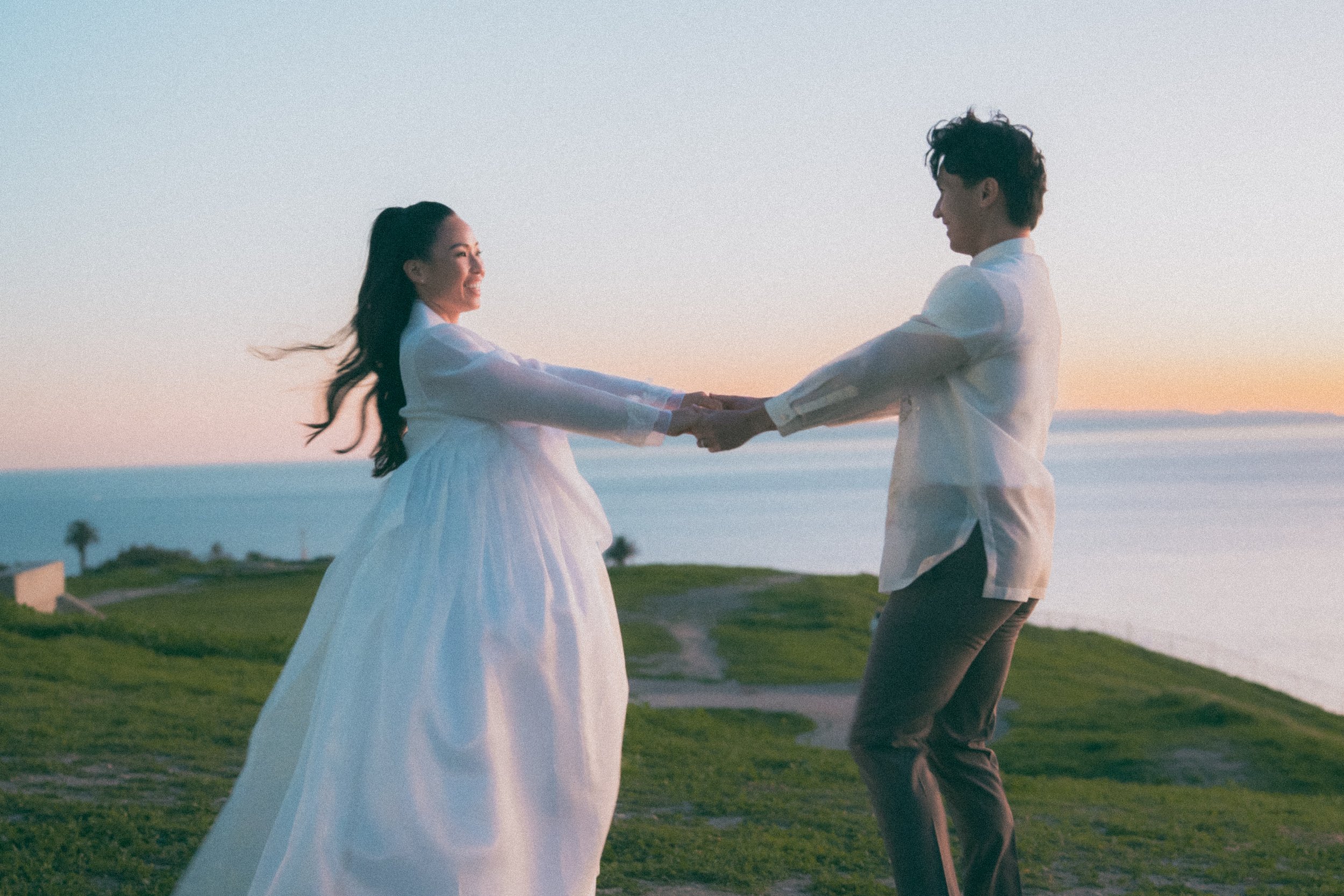 A couple holding hands and smiling at each other outdoors during sunset, with ocean and green grass in the background.