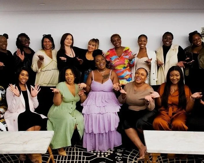 Group of women posing indoors, smiling and gesturing with their hands, dressed in various colorful outfits.