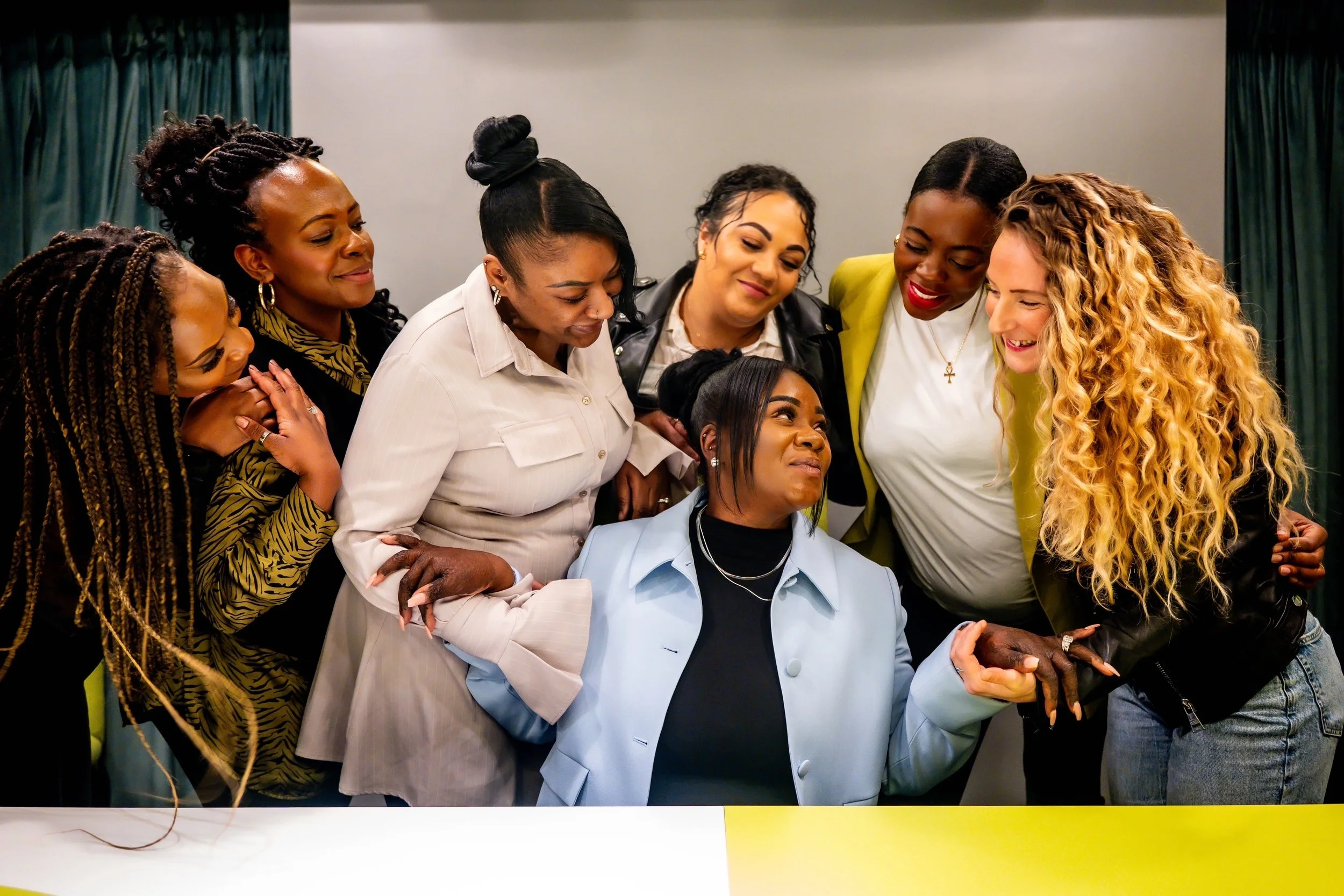 A group of seven women gathered around a seated woman, appearing to comfort and support her.