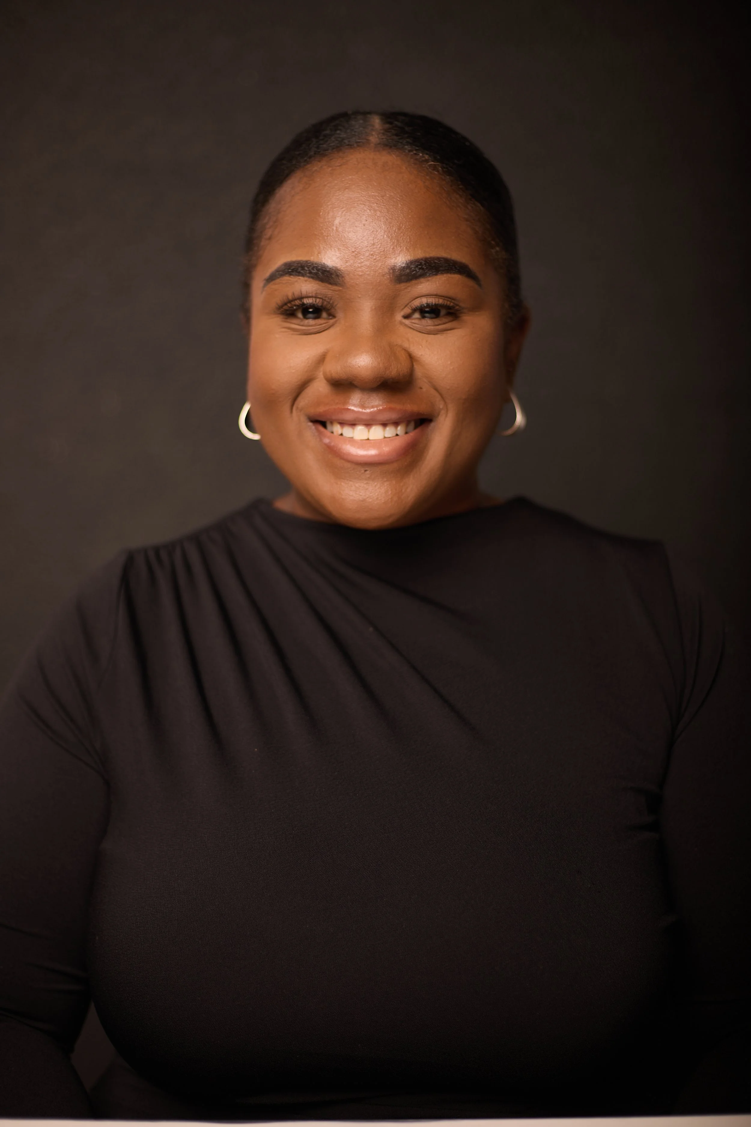 A woman with dark hair pulled back, smiling, wearing hoop earrings and a black top, against a dark background.