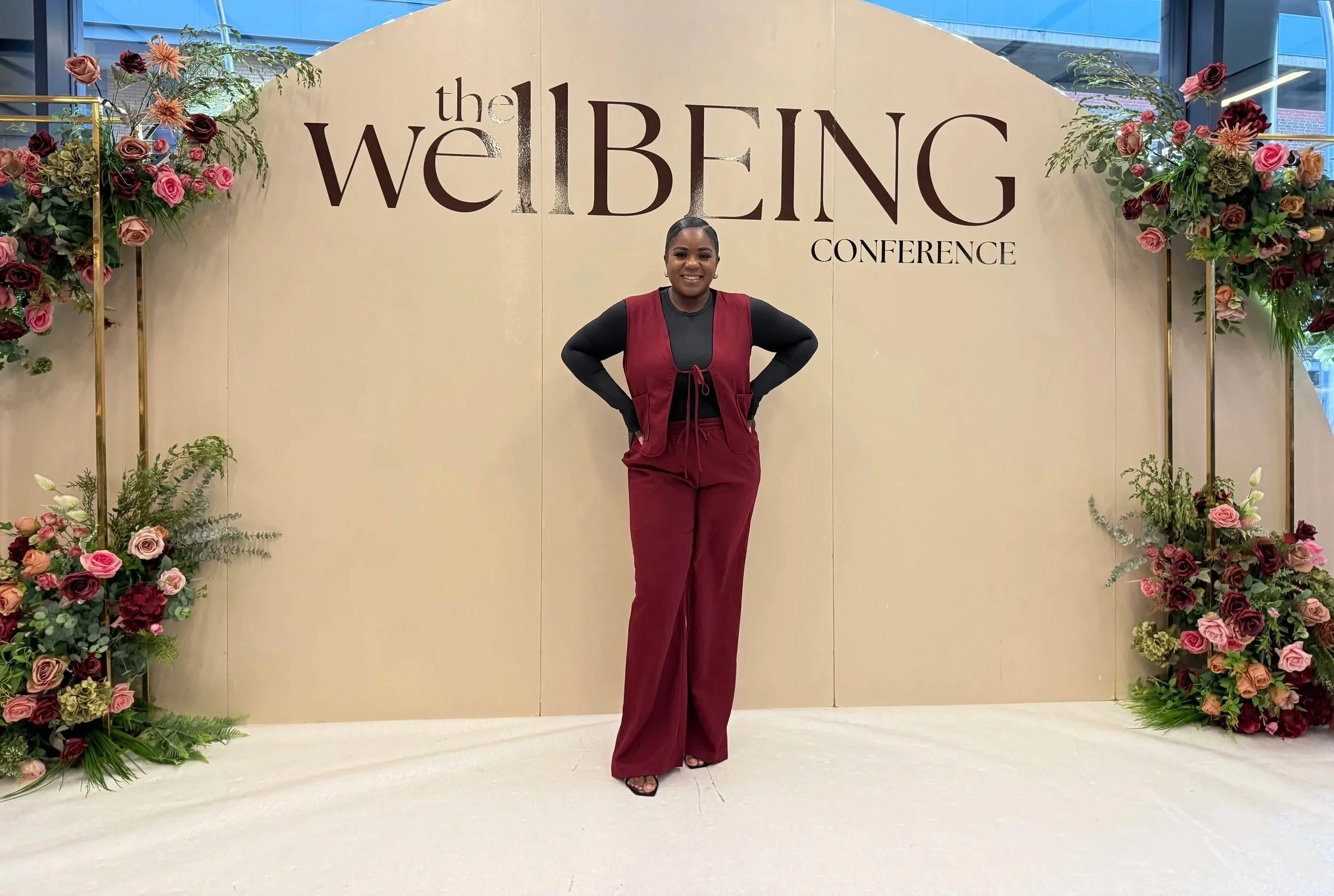 A woman standing confidently with hands on hips in front of a backdrop that reads 'the WELLBEING CONFERENCE,' surrounded by floral arrangements.
