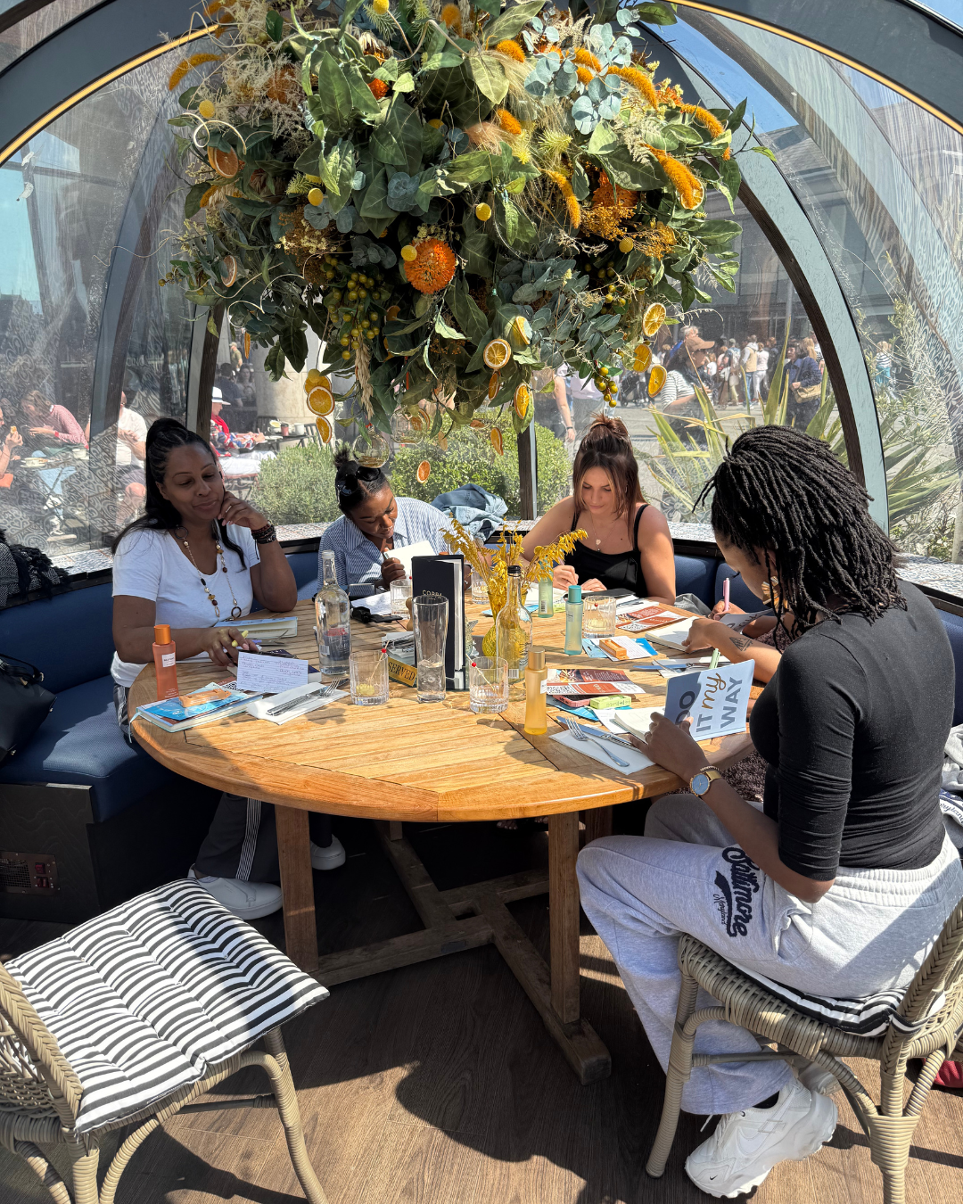 Four women sitting around a round wooden table with various drinks and papers, inside a glass-domed structure decorated with a large hanging floral arrangement with greenery and dried citrus slices.