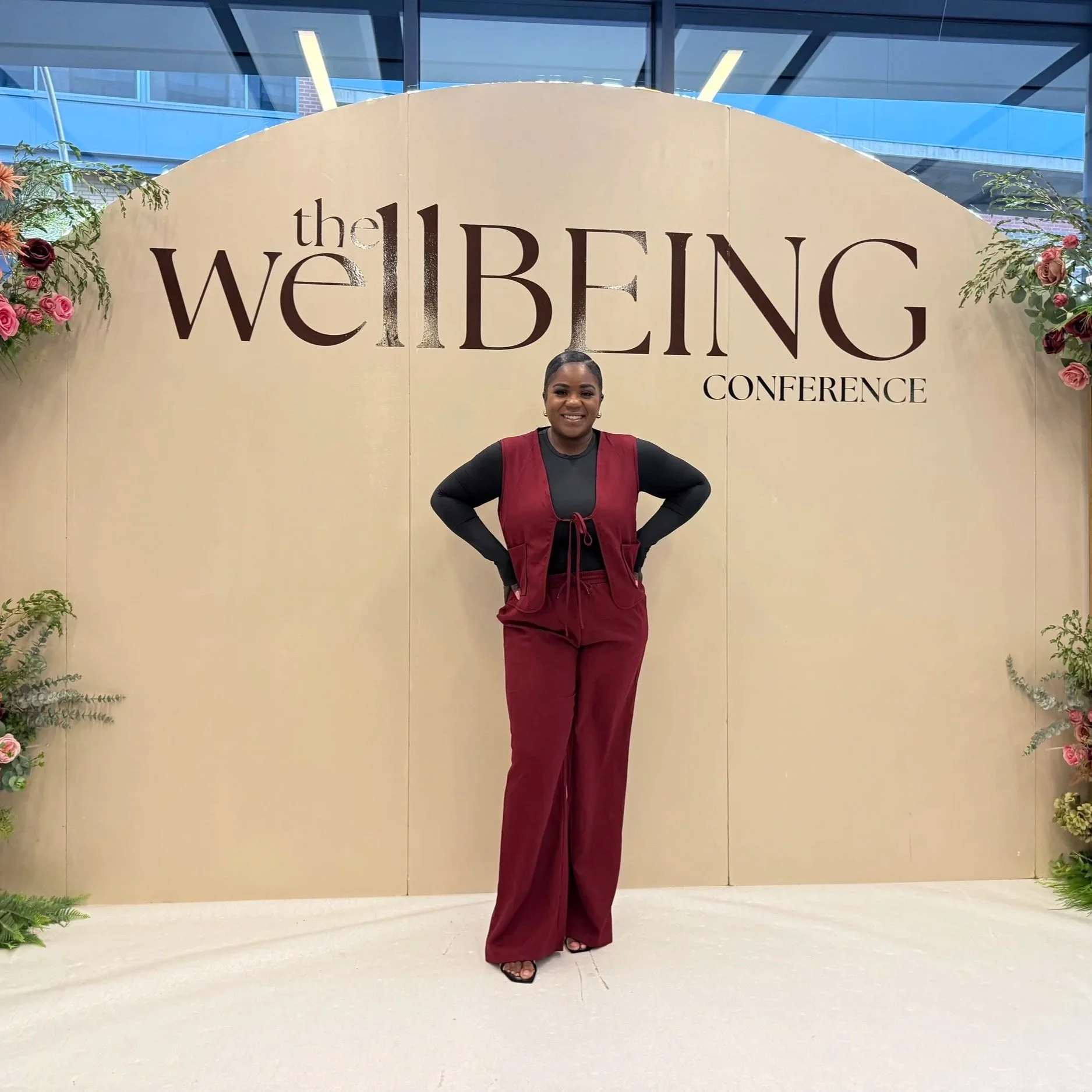 A woman standing in front of a beige backdrop with text 'the WELLBEING CONFERENCE'. She is smiling, wearing a black top, red trousers, and a matching red vest, with her hands on her hips.