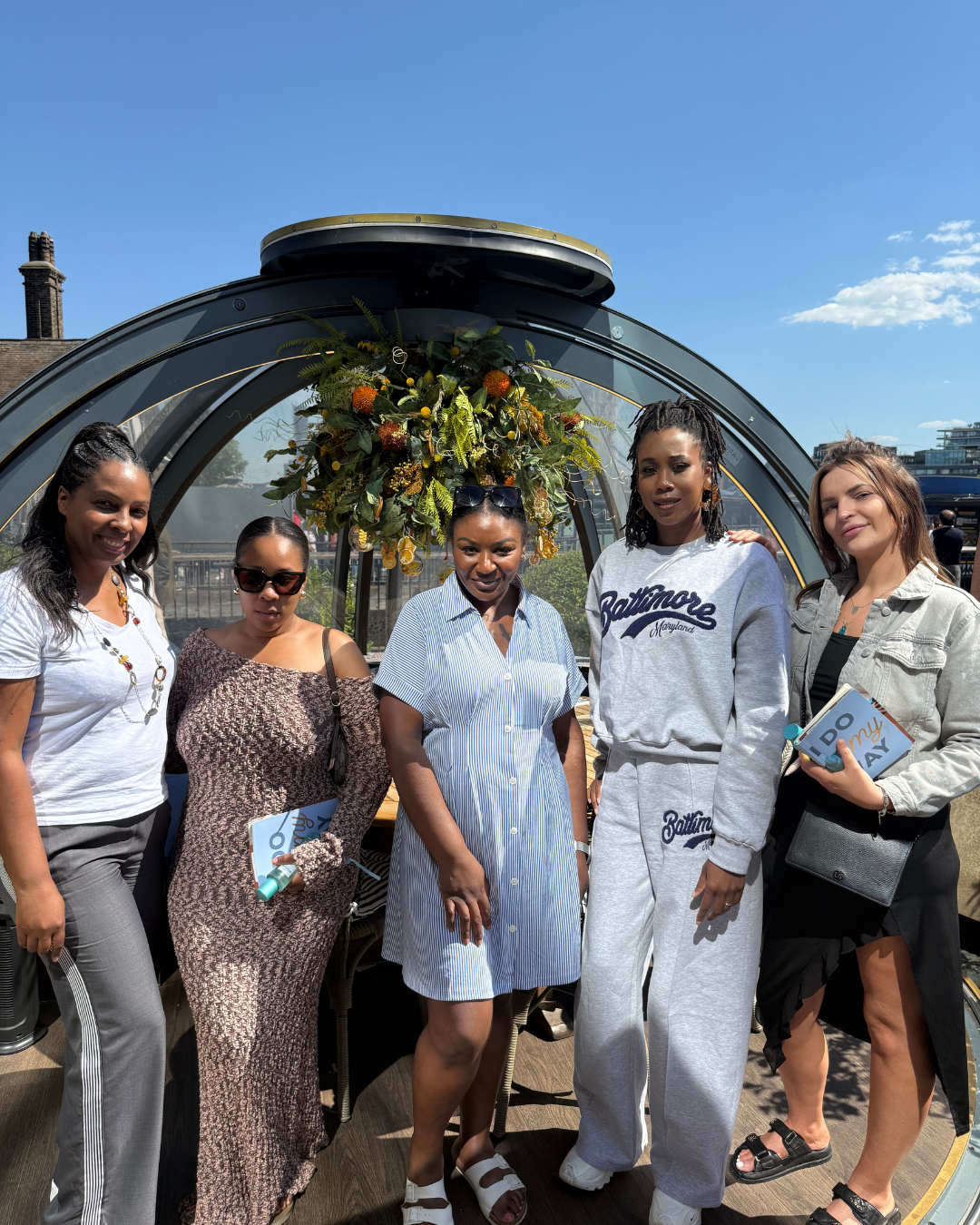 Group of five women standing outdoors in front of a structure with a floral arrangement on top, smiling at the camera on a sunny day.