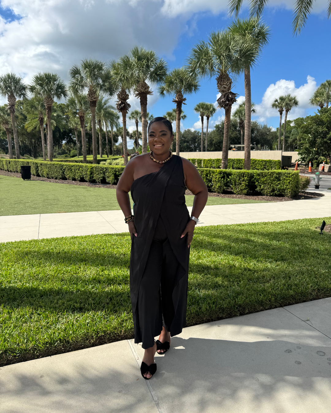 Woman standing outdoors on a sidewalk in front of palm trees, wearing a black one-shoulder dress and jewelry, smiling.