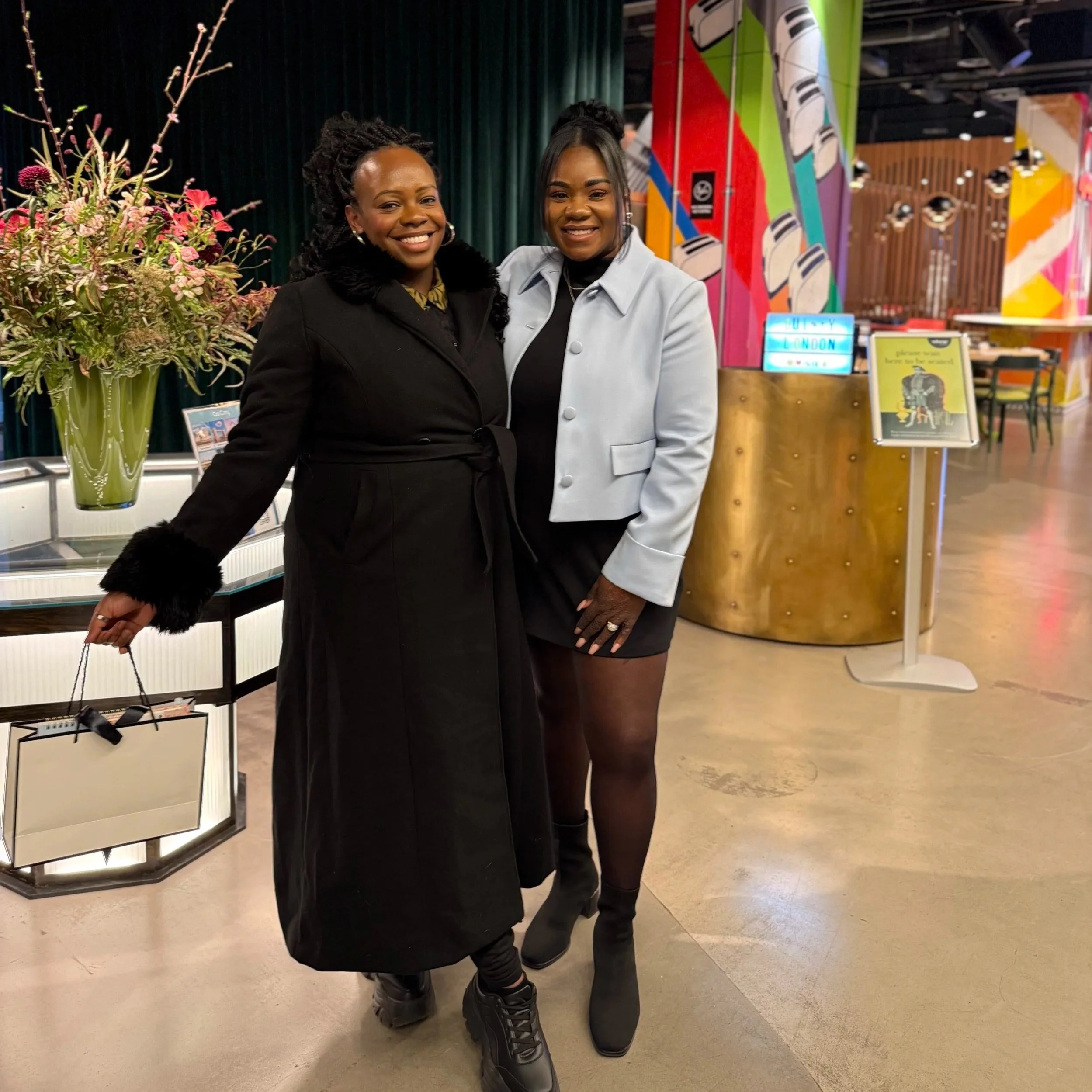 Two women standing together inside a colorful venue, smiling for the photo, with a large floral arrangement and colorful abstract art in the background.