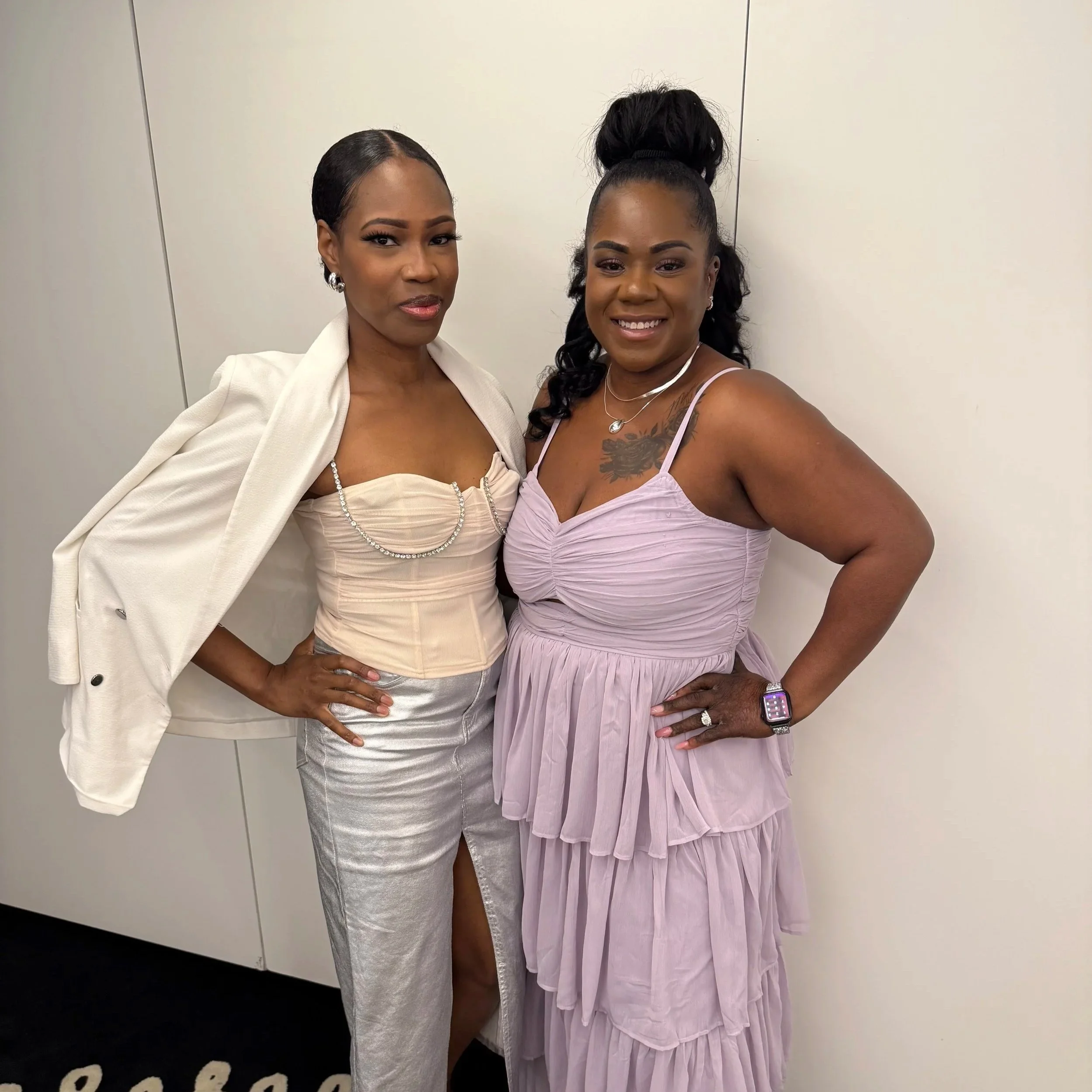 Two women posing together indoors, both wearing elegant dresses and jewelry, smiling at the camera.