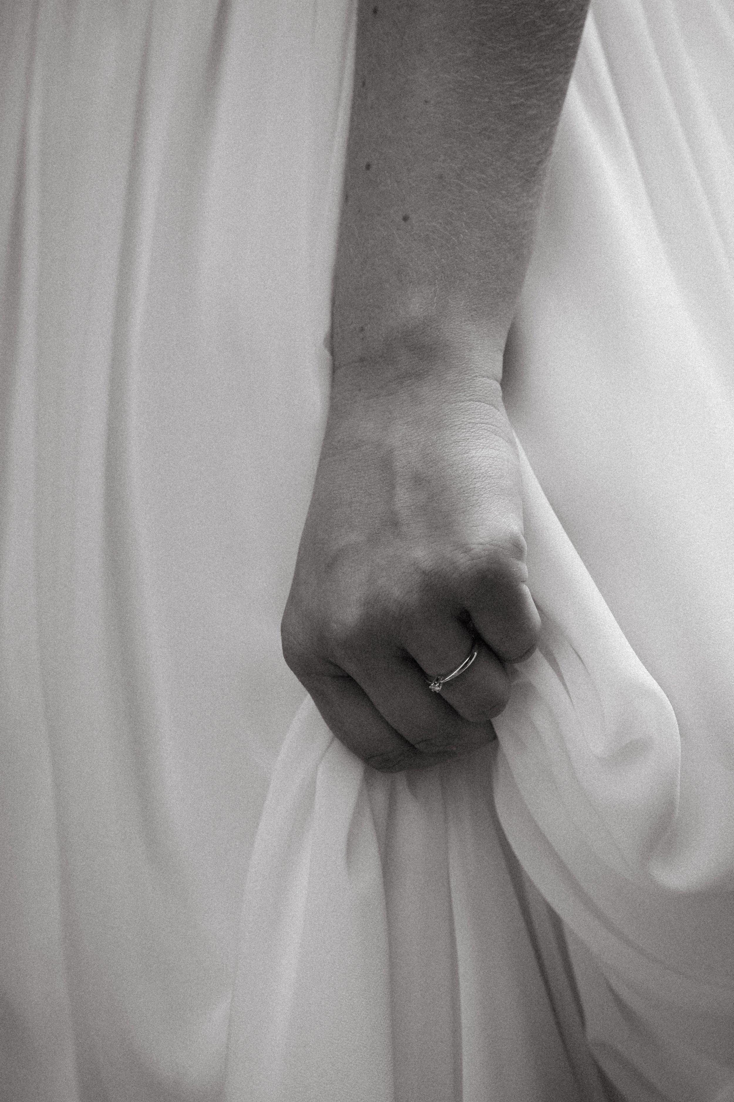Nahaufnahme einer Hand, die ein weißes Kleid hält, mit einem Verlobungsring am Finger.