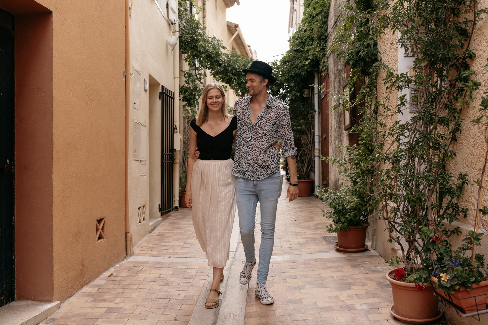Ein lächelndes Paar spaziert Arm in Arm durch eine malerische, enge Gasse, umgeben von Pflanzen und Gebäuden mit mediterranen Farben.