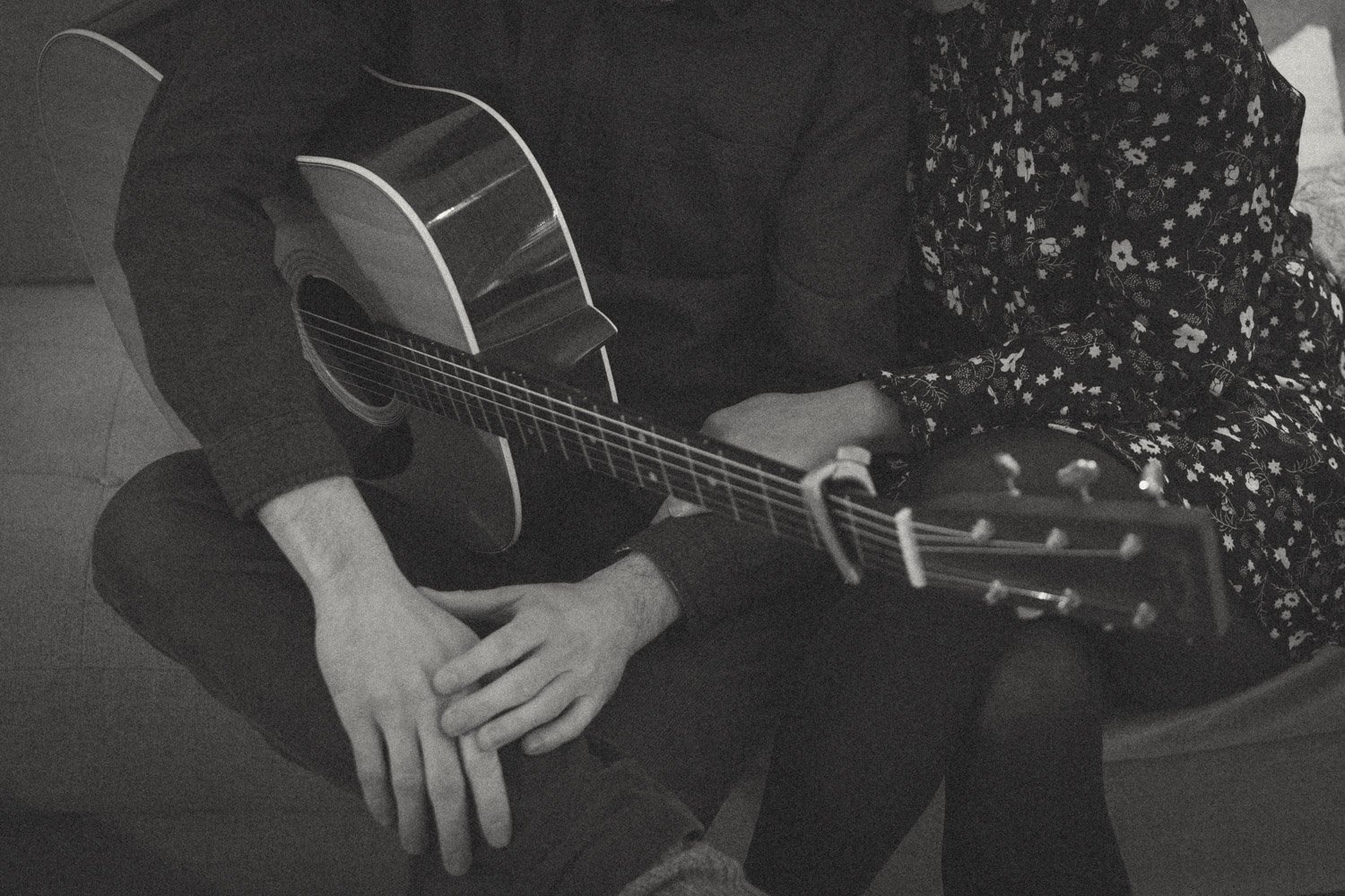 Zwei Personen auf einem Sofa mit einer Akustikgitarre, schwarz-weiß Bild. Eine Person trägt ein florales Kleid, während die andere lässige Kleidung trägt. Die Gitarre liegt auf den Knien beider Personen.