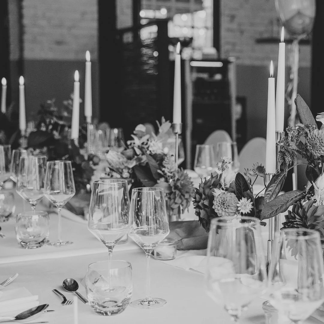 Schwarz-weiß-Foto eines elegant gedeckten Tisches mit Weingläsern, Besteck, Kerzen und Blumenarrangements.