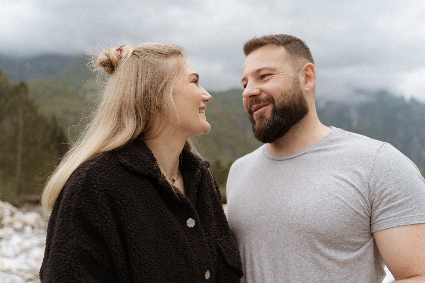 Ein Paar lächelt sich vor einem Berg- und Waldhintergrund an. Die Frau hat blondes Haar und trägt eine dunkle Jacke, der Mann hat einen Bart und trägt ein graues T-Shirt.