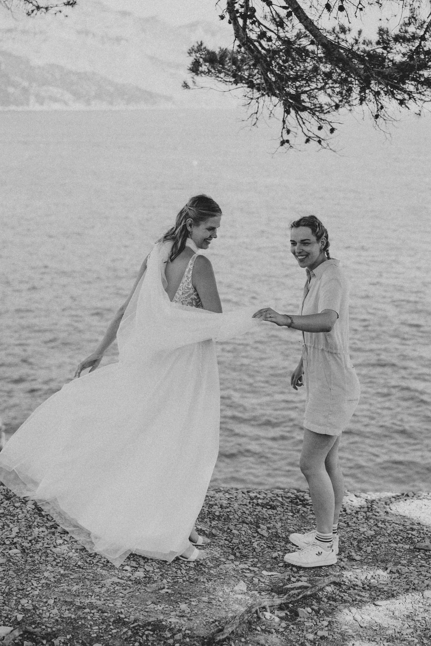 Zwei Personen lächeln am Ufer mit Wasser im Hintergrund. Eine Person trägt ein langes, fließendes Kleid; die andere trägt einen Jumpsuit und Sneakers. Es ist ein schwarz-weißes Bild.