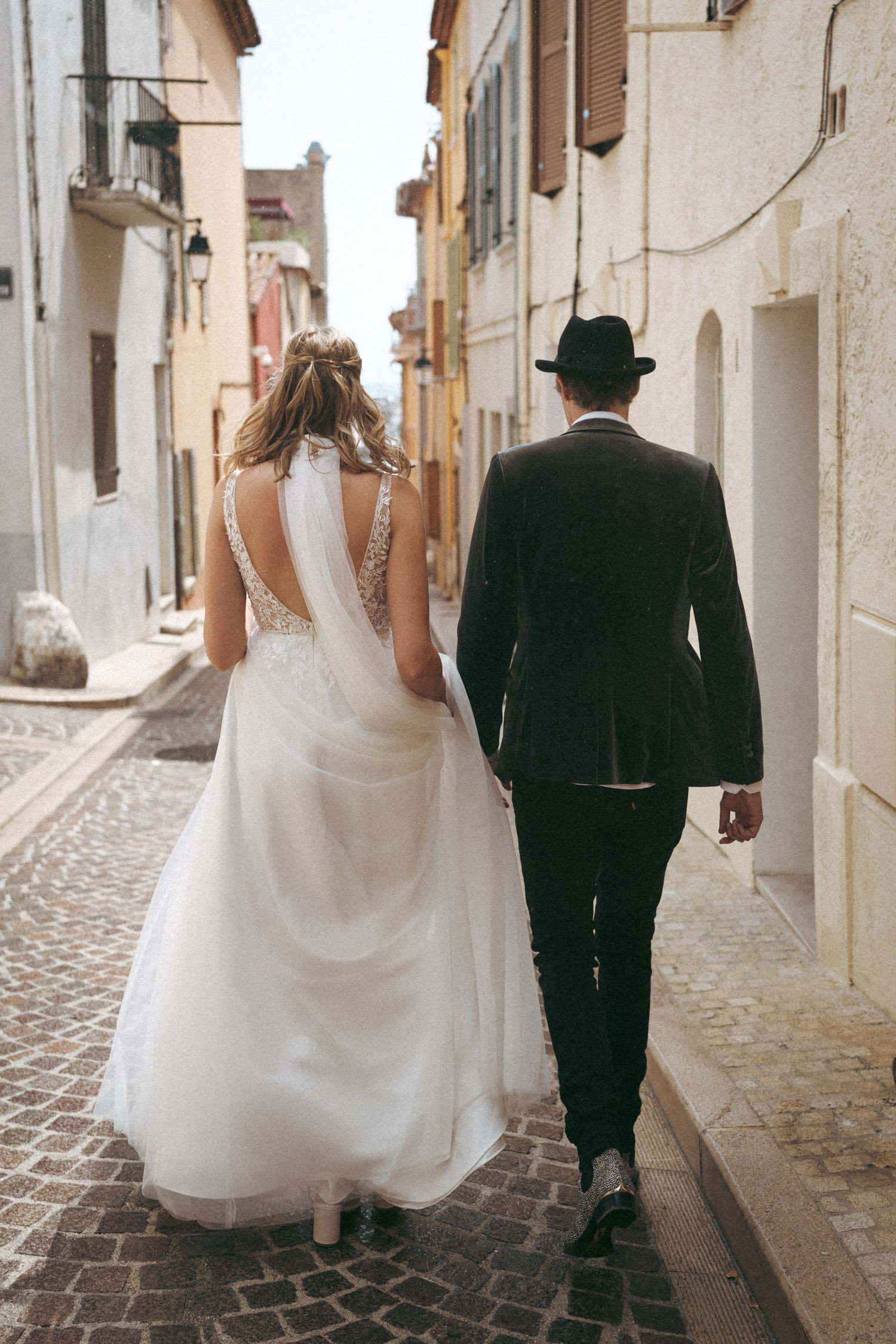 Ein Brautpaar geht eine gepflasterte Straße entlang, umgeben von Gebäuden. Die Braut trägt ein weißes Hochzeitskleid und der Bräutigam ist im Anzug gekleidet.