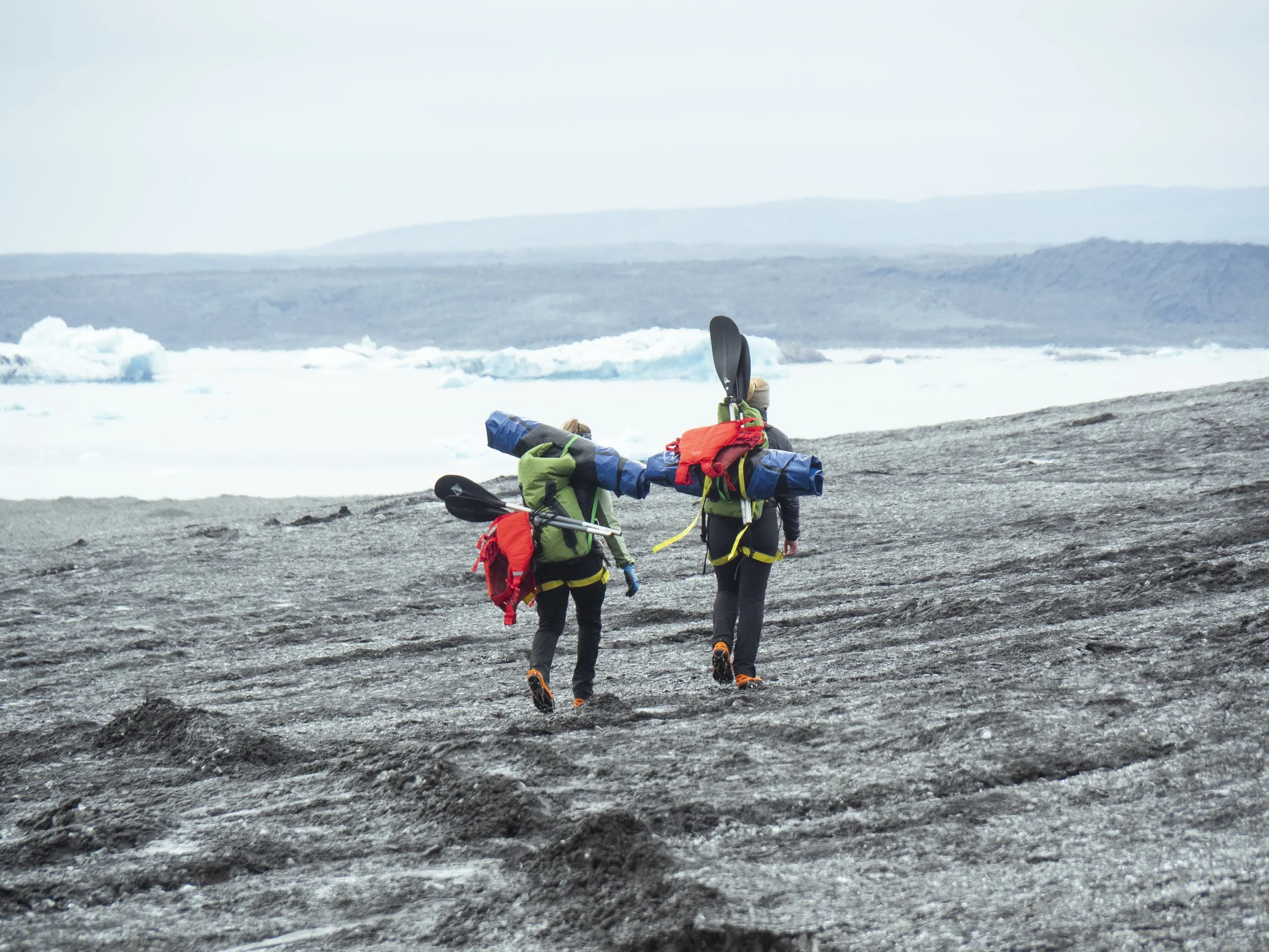 Two hikers carrying camping gear walk on rocky terrain near a partially icy body of water in a cold, remote landscape.