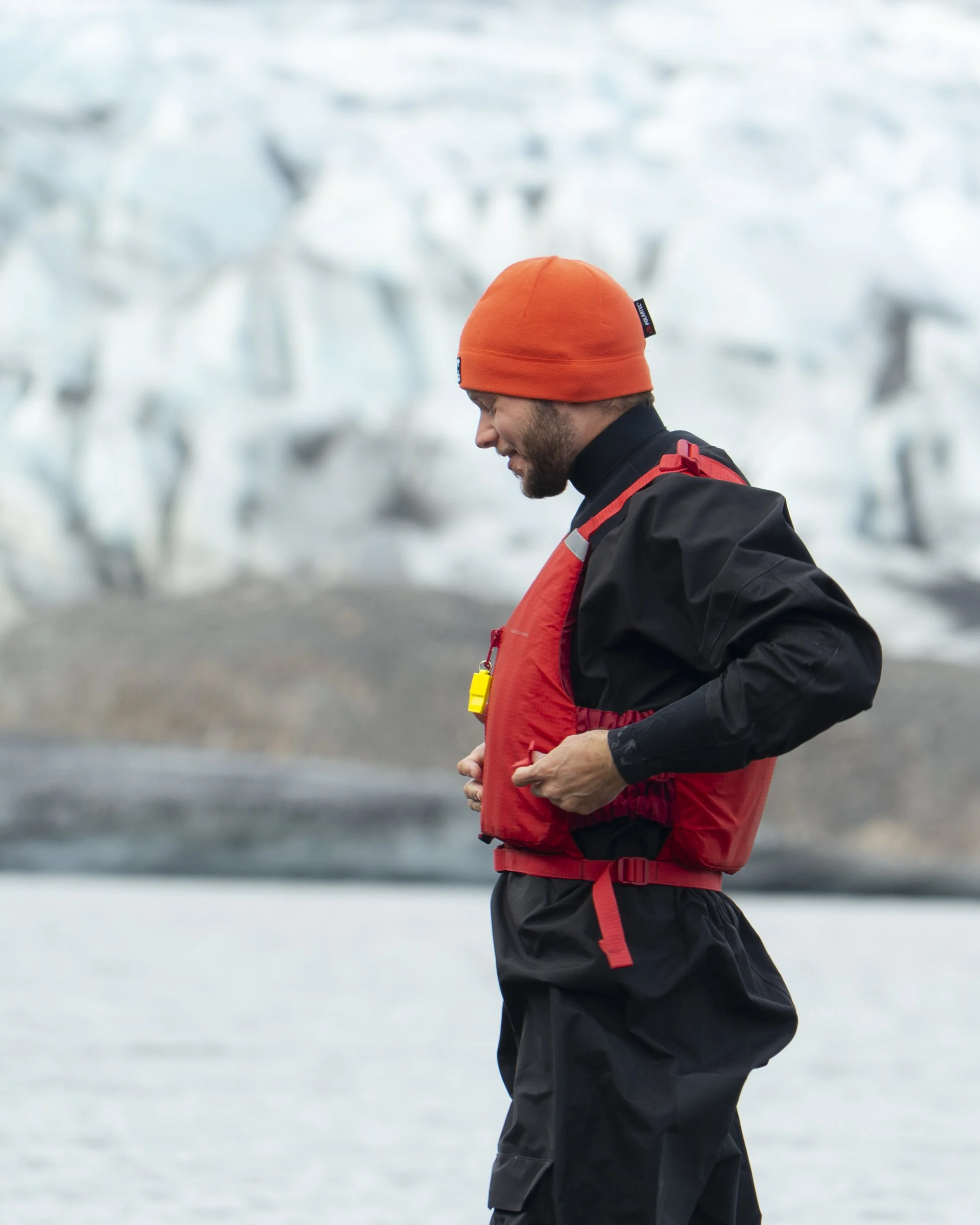A man in black outdoor gear with an orange beanie and red life vest stands on a cold, icy landscape with icebergs in the background.