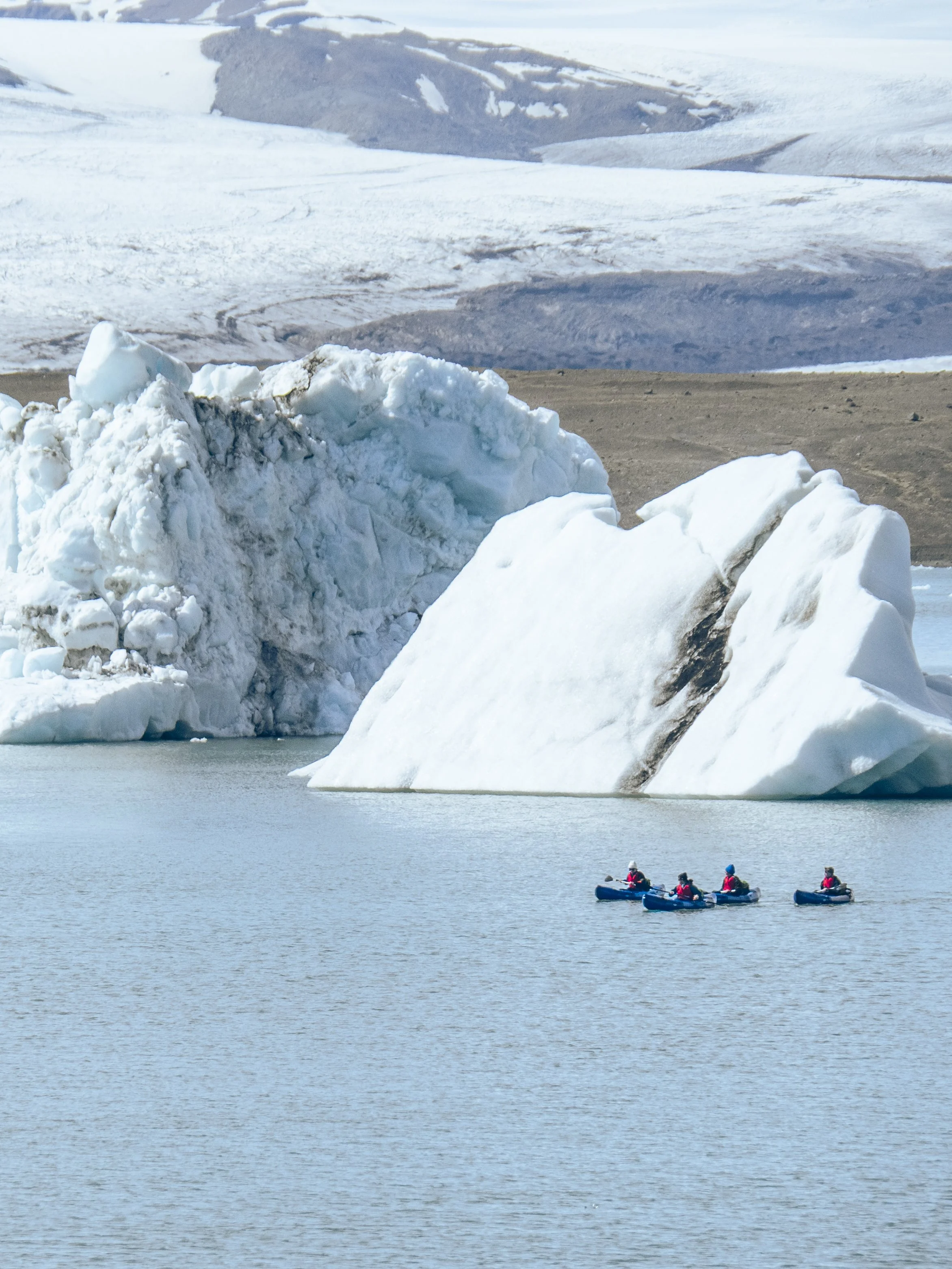 Four people in kayaks on water near large icebergs and a snowy landscape.