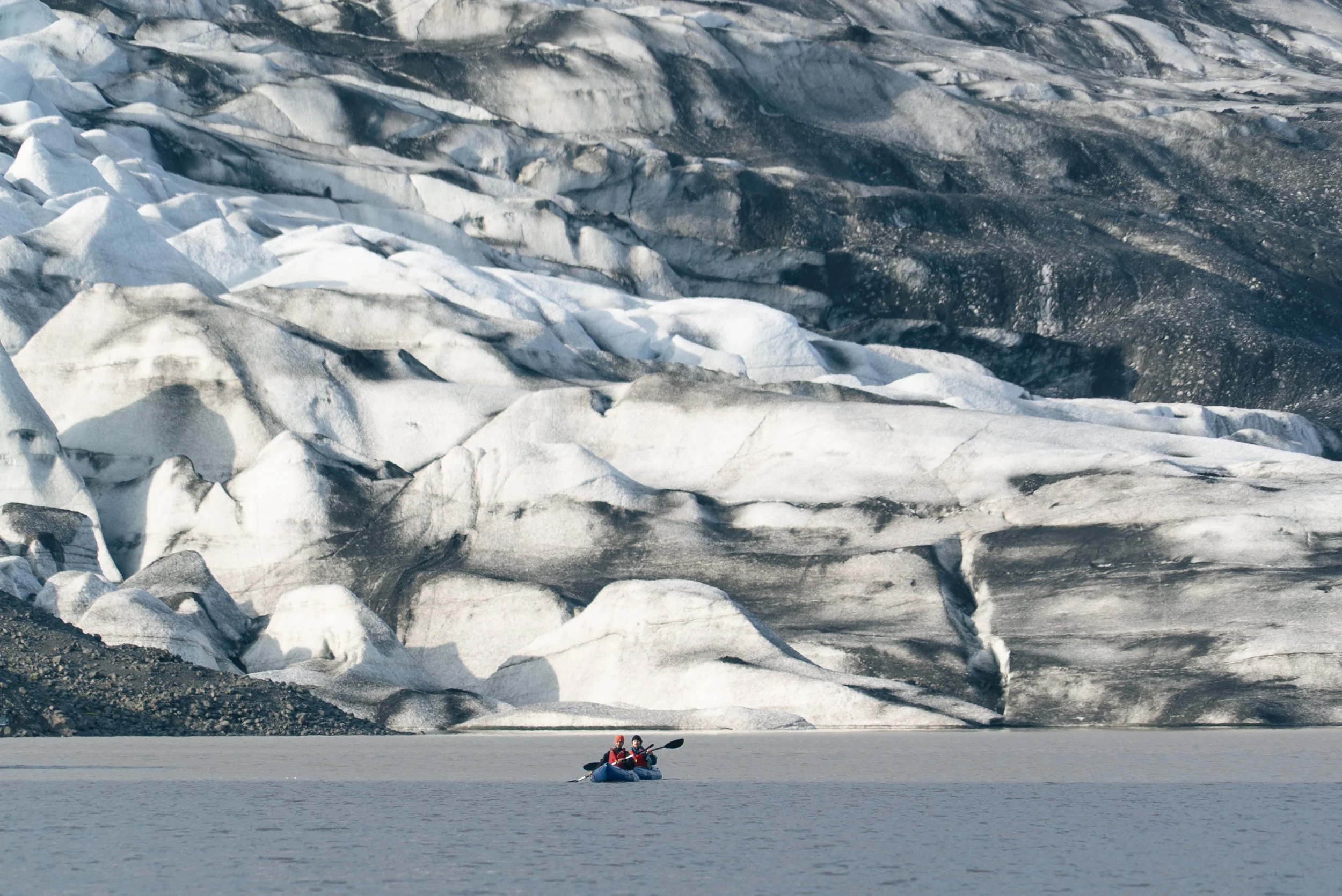 Two people in a kayak paddling near a glacier with large ice formations and dark rocky terrain in the background.