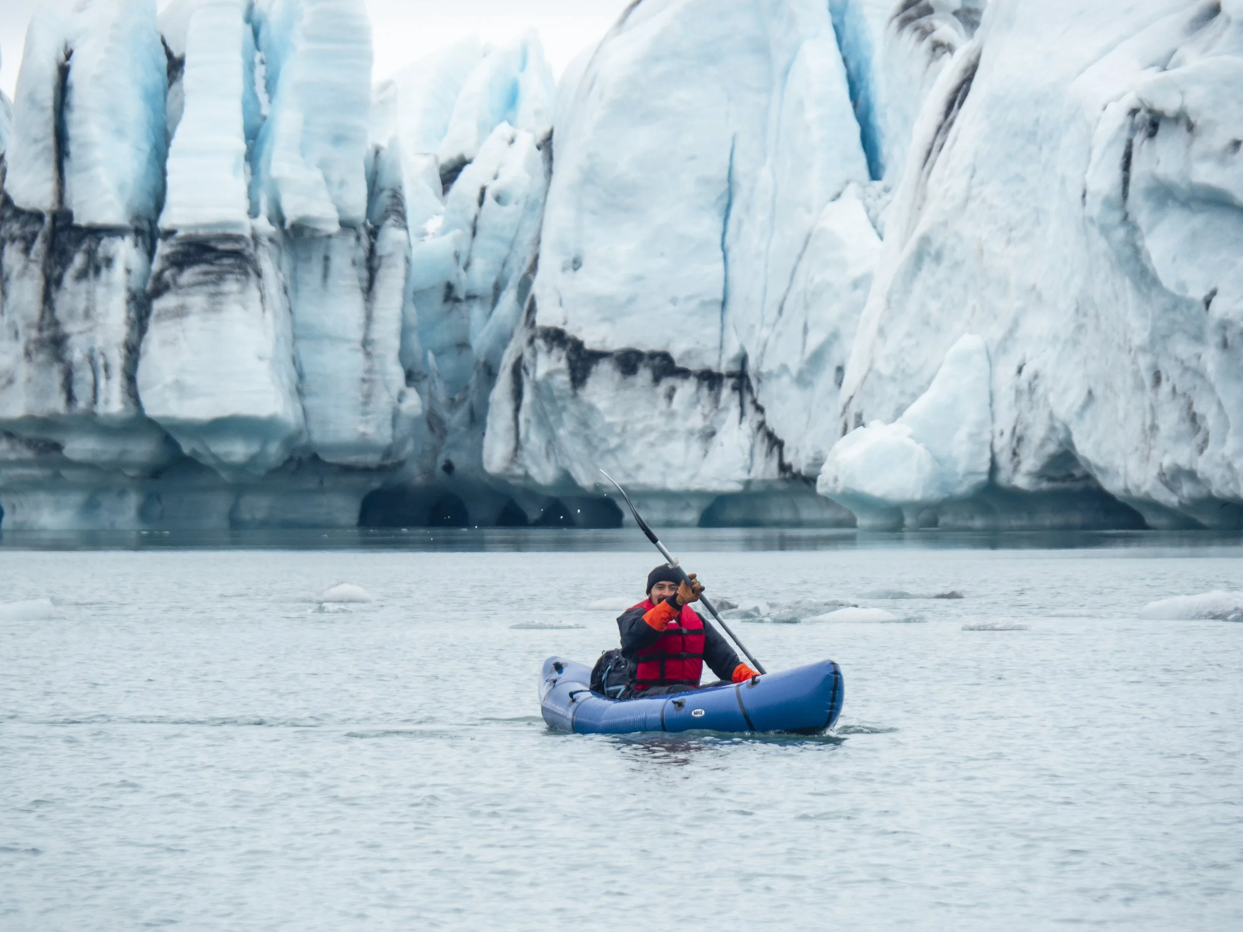 Person in a blue kayak paddling in icy water with icebergs and a glacier in the background.