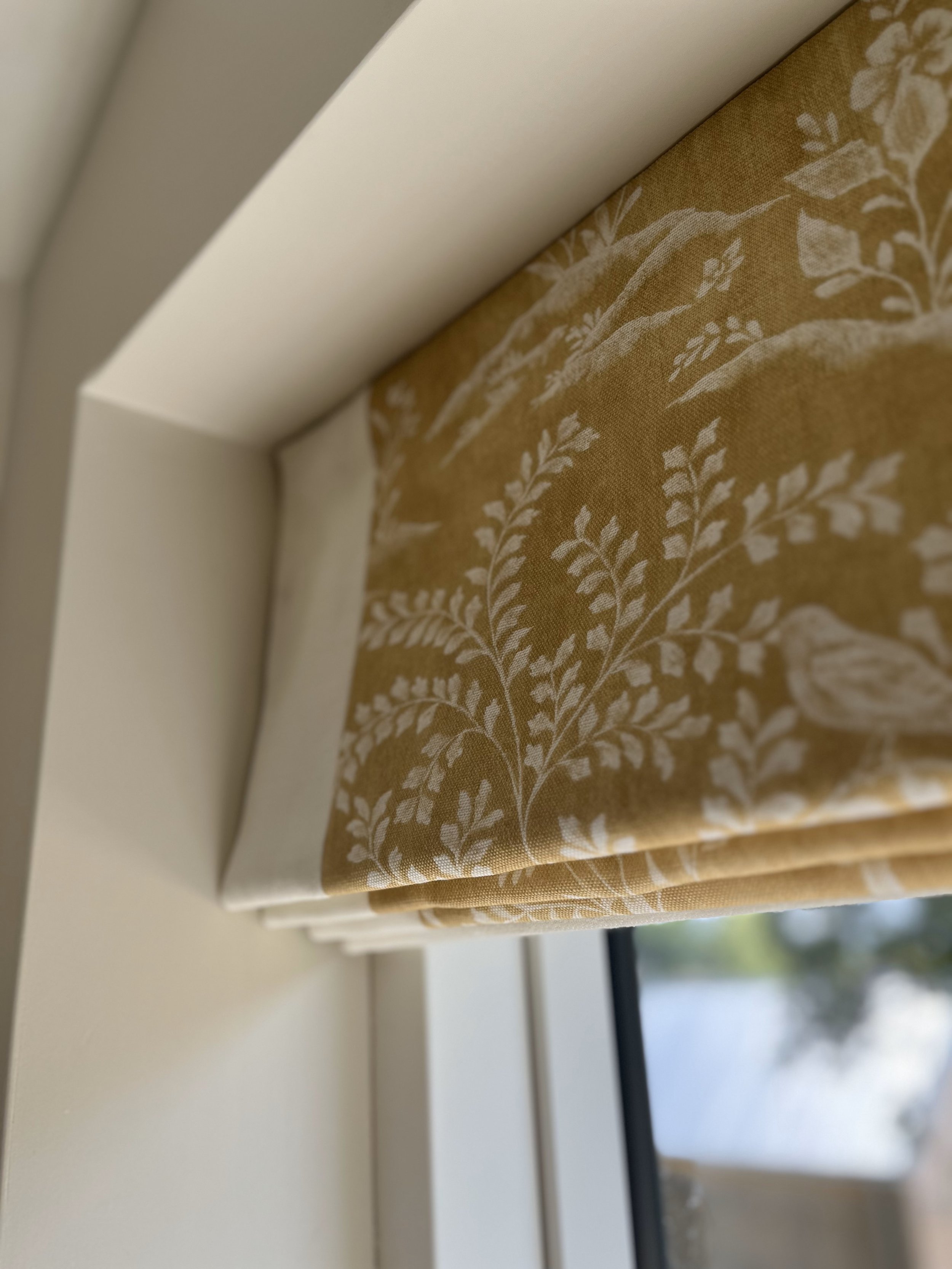 Close-up of a window with a folded, patterned yellow Roman shade featuring white leaf and bird designs.