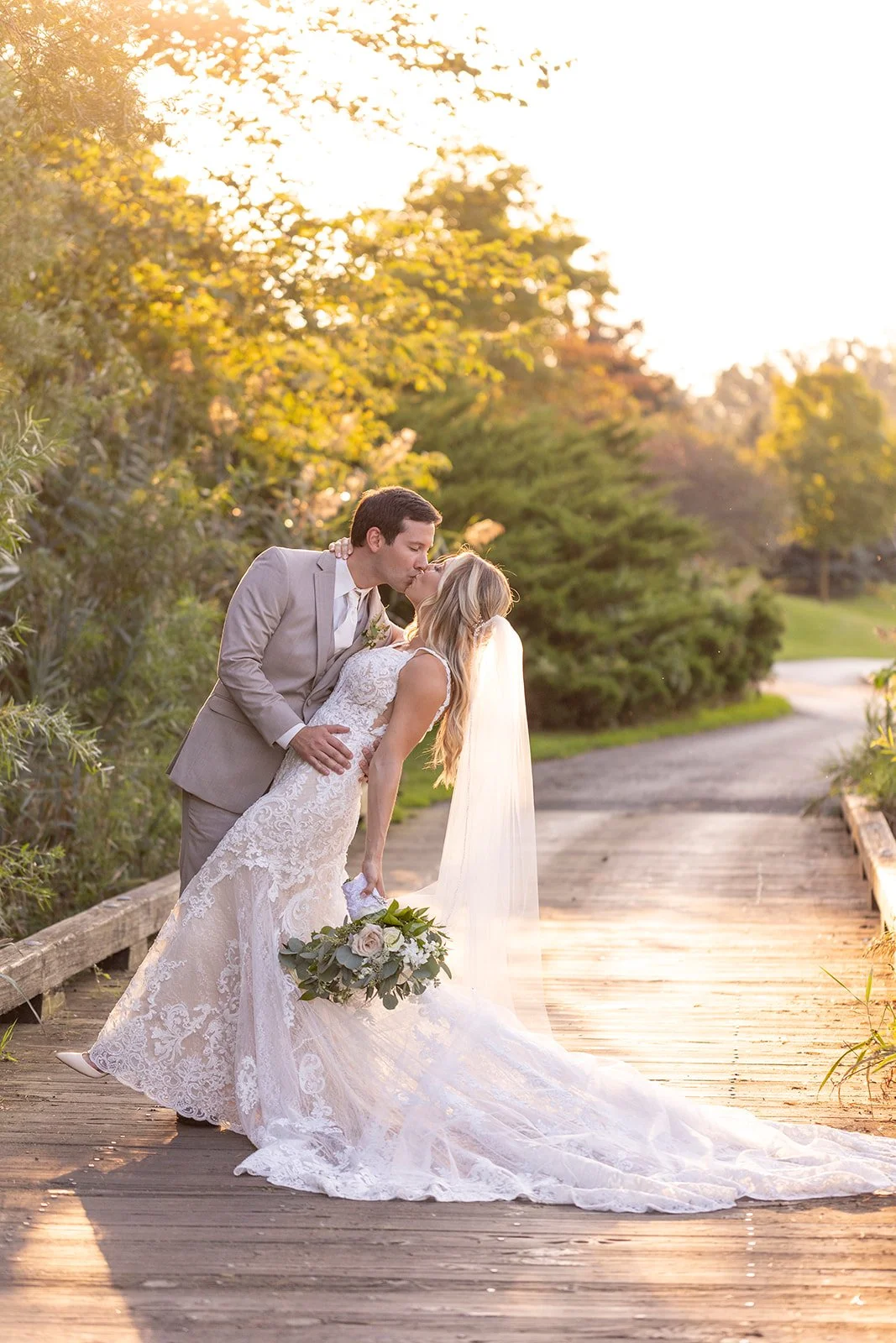 A bride and groom share a kiss on a wooden bridge during sunset, surrounded by greenery, with the bride holding a bouquet of flowers.