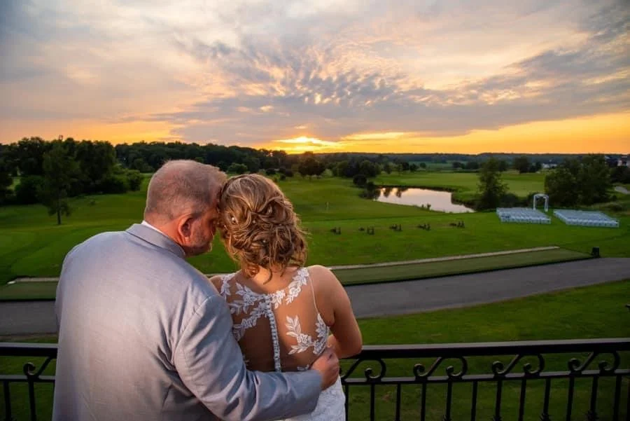 A couple stands closely together on a balcony overlooking a green landscape at sunset, with rolling hills, a pond, and a partly cloudy sky in the background.