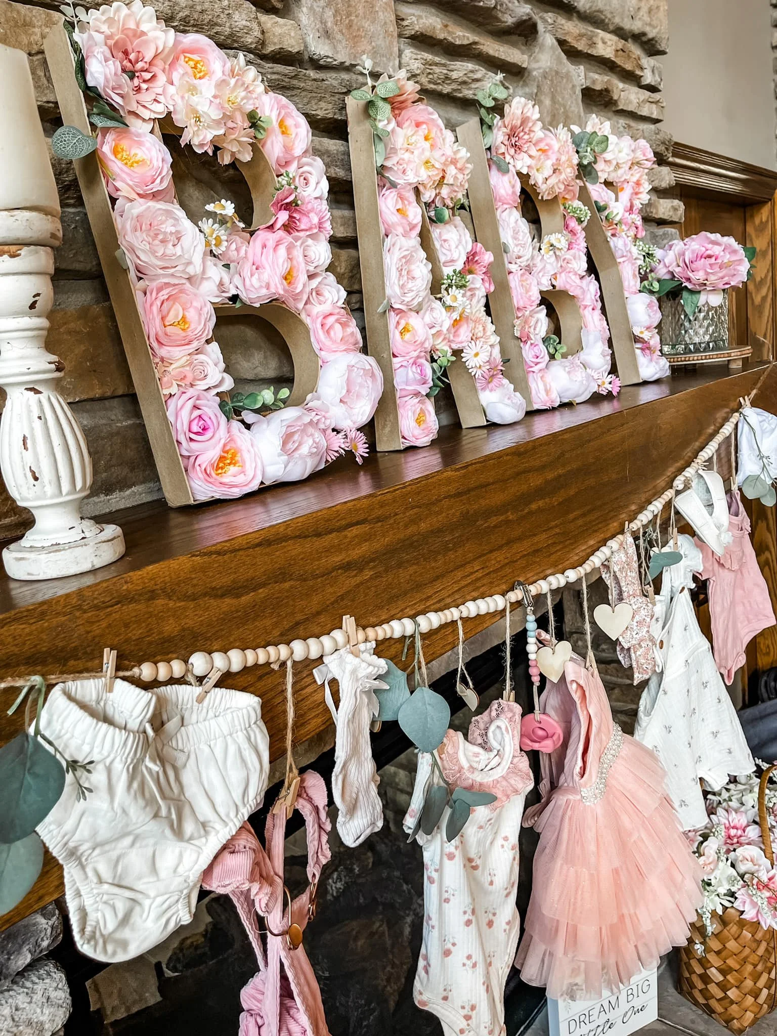 Decorative fireplace mantel with large wooden letters spelling 'BABY' adorned with pink and white flowers, and a clothesline with baby clothing and accessories hanging underneath.