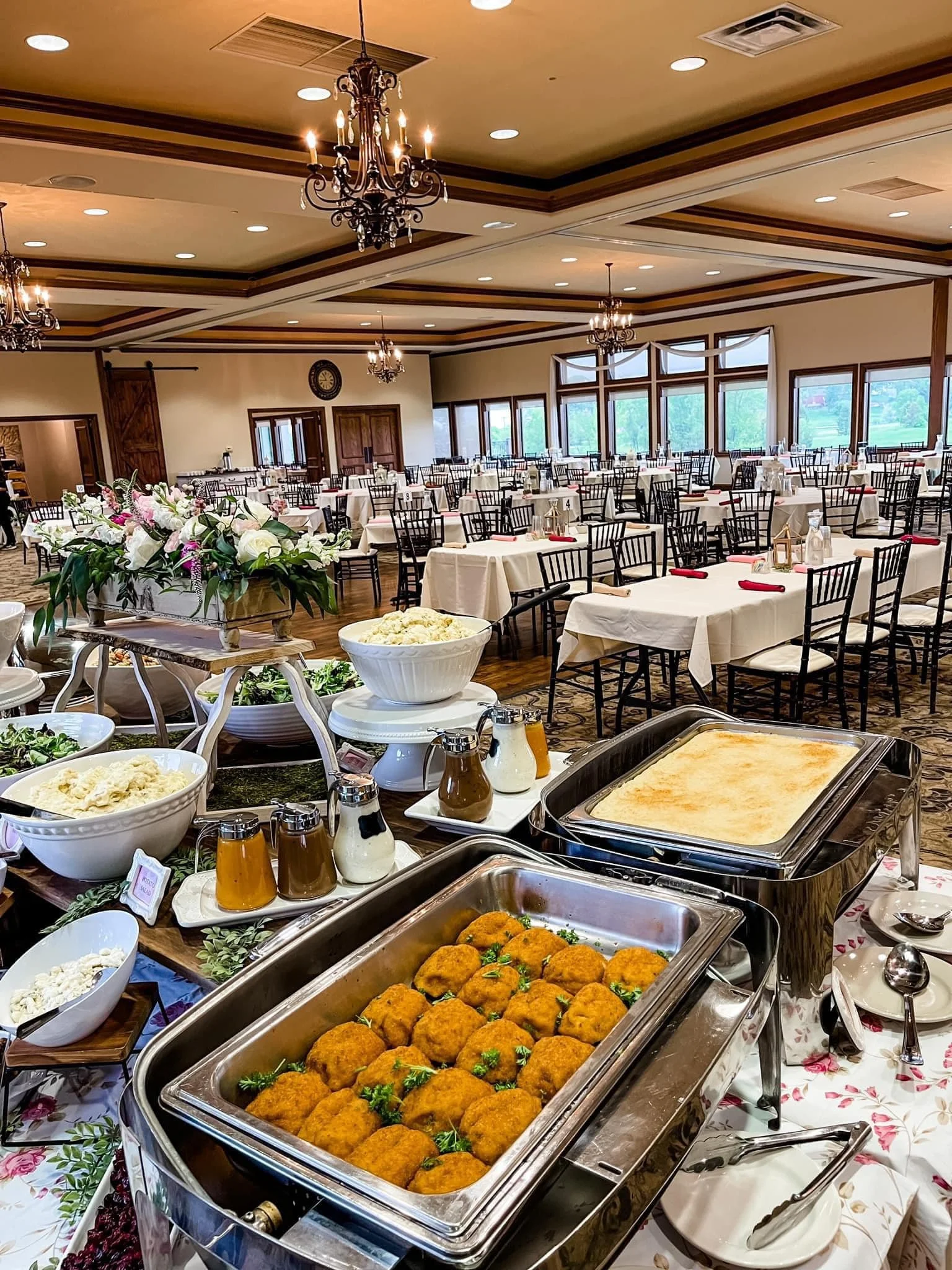 Buffet table with fried chicken and baked dish, set in a banquet hall with round tables, floral arrangements, chandeliers, and large windows.