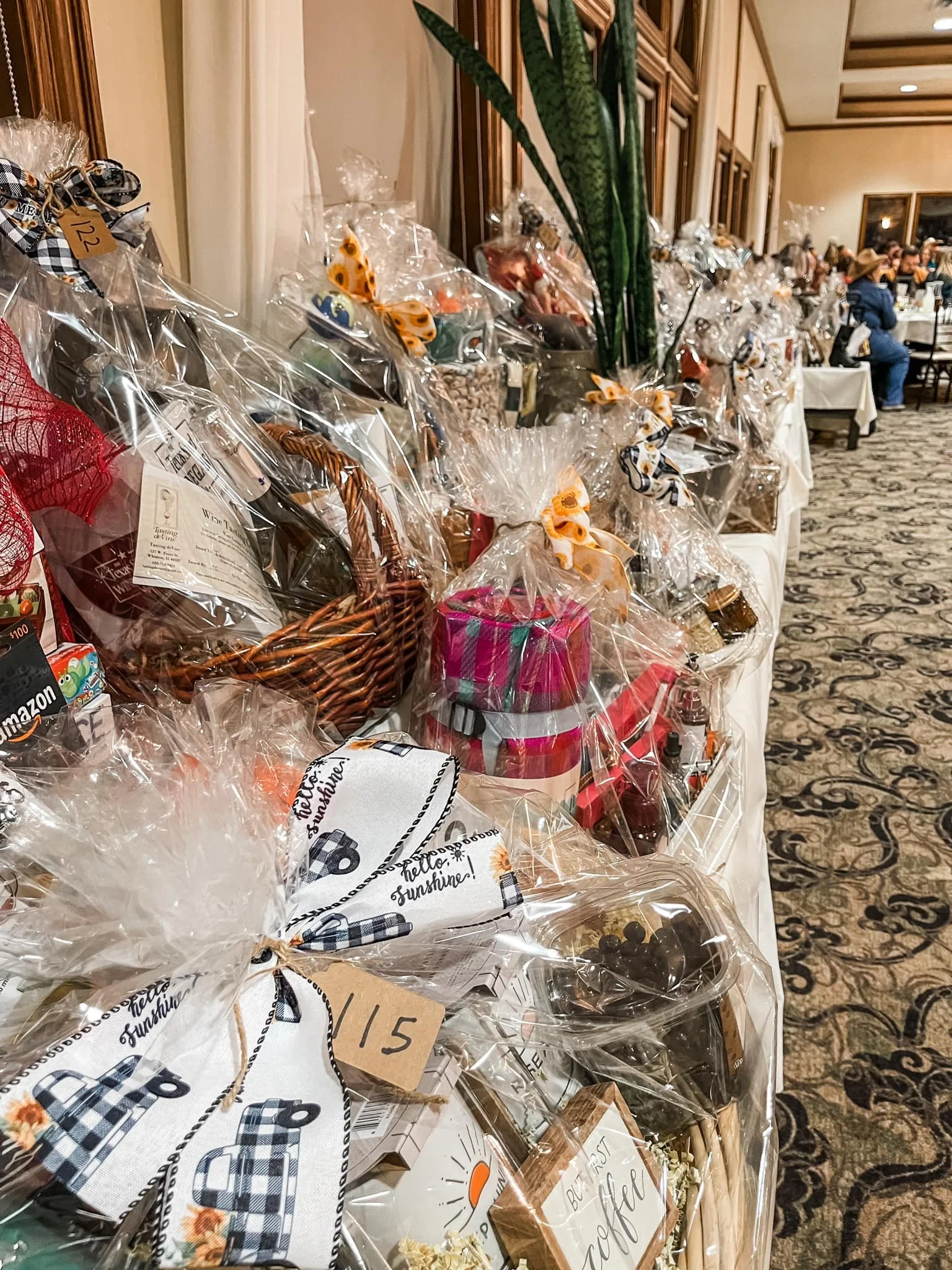 Table displaying numerous gift baskets wrapped in clear plastic with ribbons, located in a banquet hall with people seated in the background.