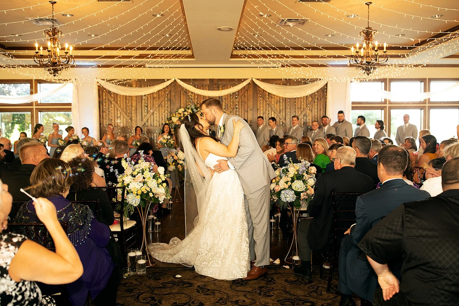 A newlywed couple sharing a kiss at the altar during a wedding ceremony, with guests seated around and a wedding party in the background, inside a decorated venue with string lights and floral arrangements.