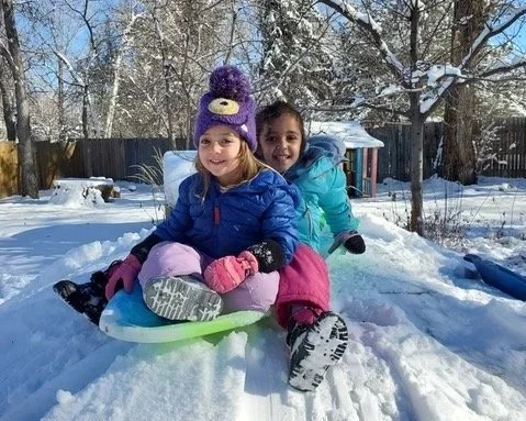 Two young girls sitting on a sled in the snow, smiling, with trees and a fence in the background, in a winter landscape.