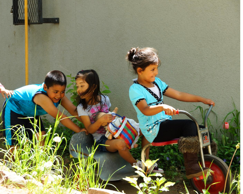 Three children playing outside, one girl riding a tricycle, and two children helping the girl on the tricycle.
