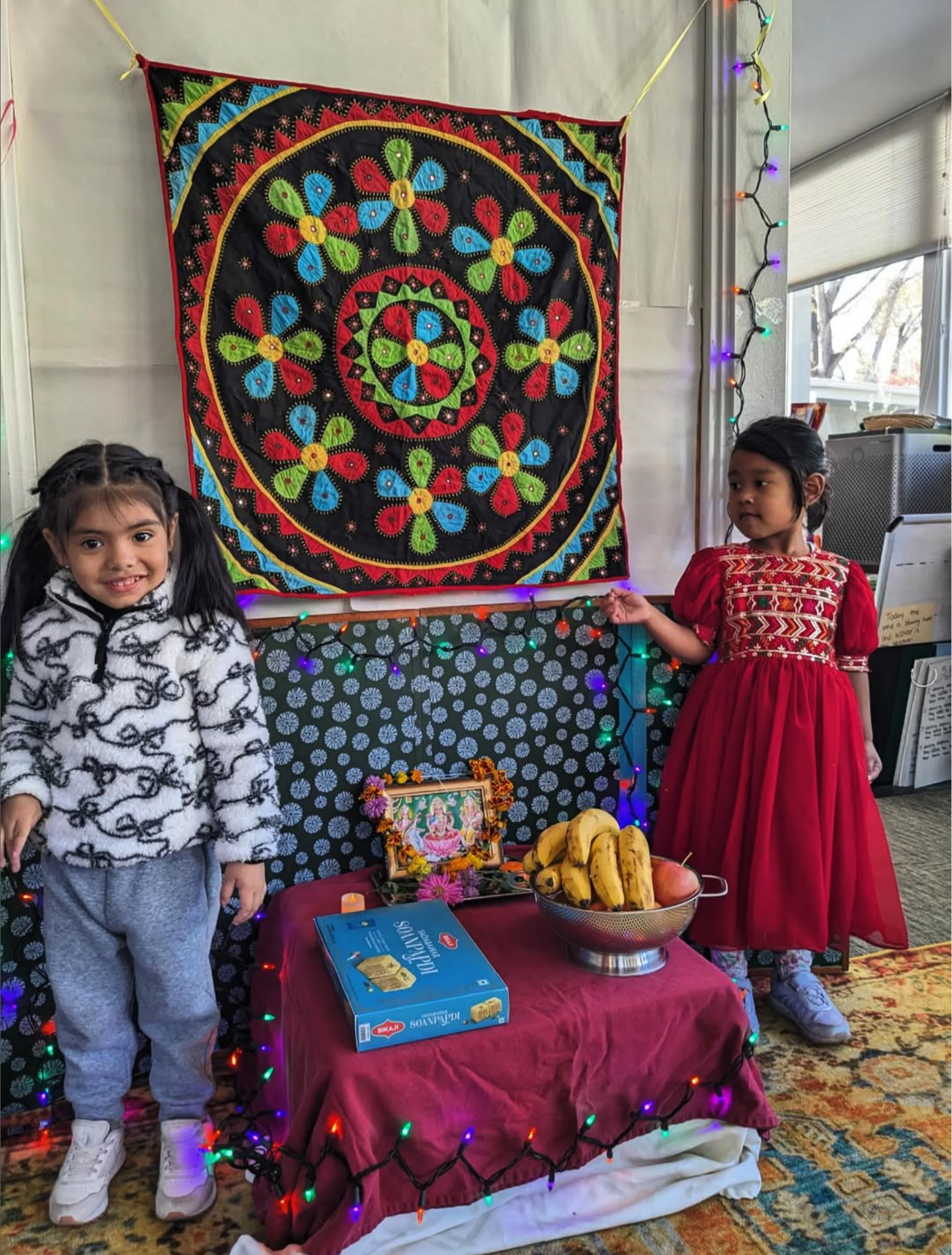 Two young girls, one wearing a black and white patterned hoodie and gray sweatpants, the other in a red dress with gold embroidery, standing beside a decorated table with bananas and a box of crackers, in front of a colorful mandala textile wall hanging with string lights.