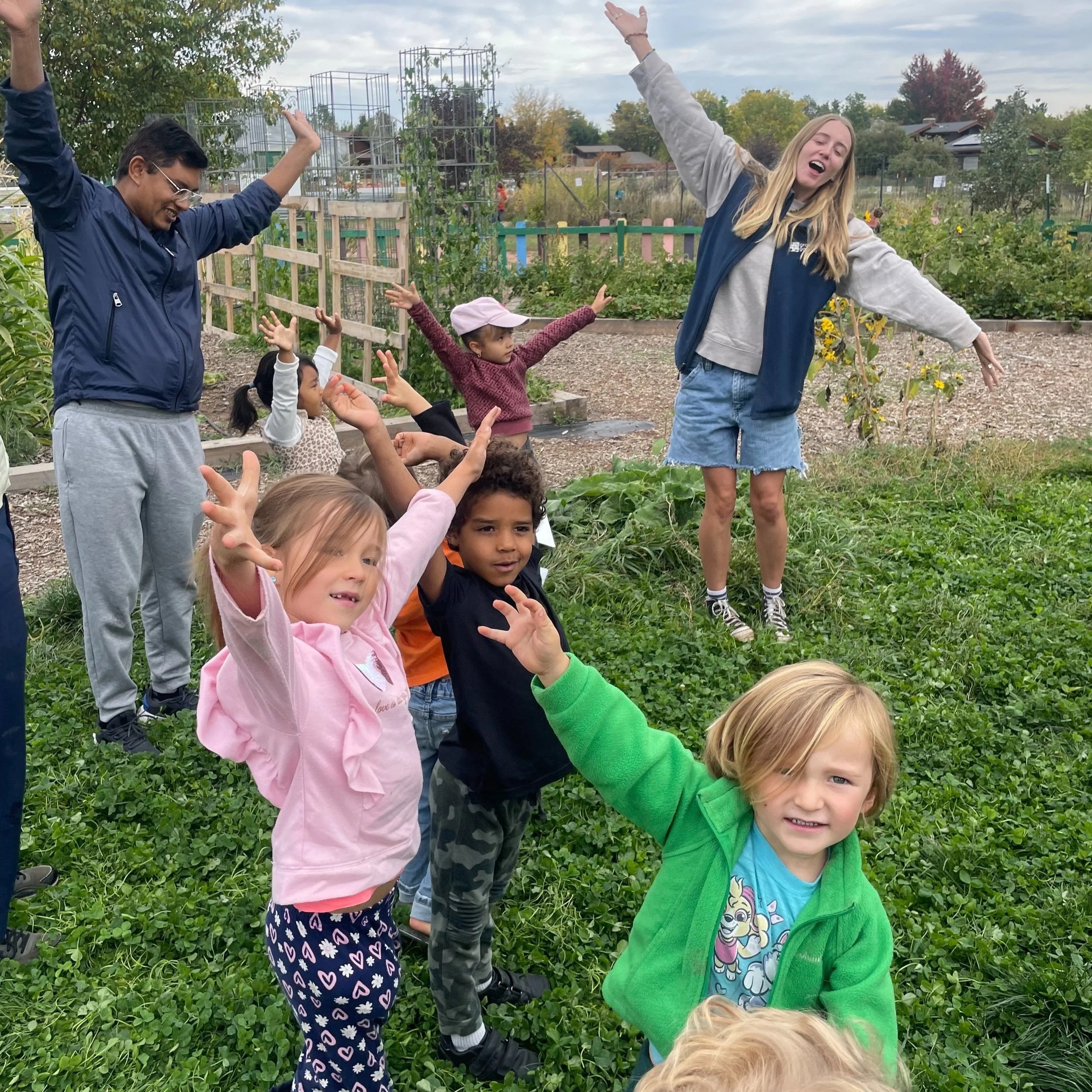 A group of children and two women in a garden with green plants and a colorful fence in the background. The children and women are happily raising their arms and smiling.