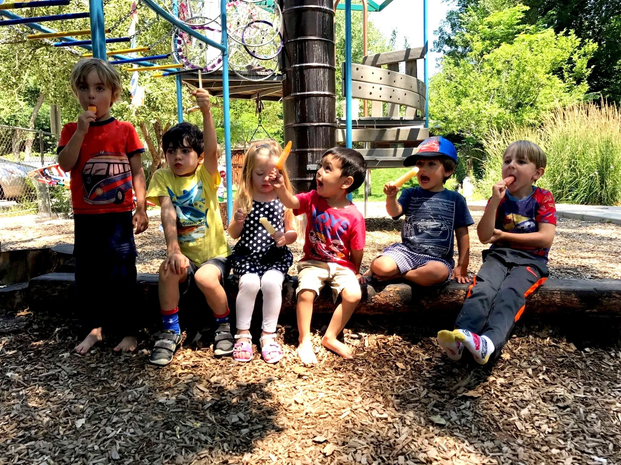 Seven children of various ethnicities sitting and standing on wood chips at a playground, holding carrots and eating snacks, with playground equipment and trees in the background.