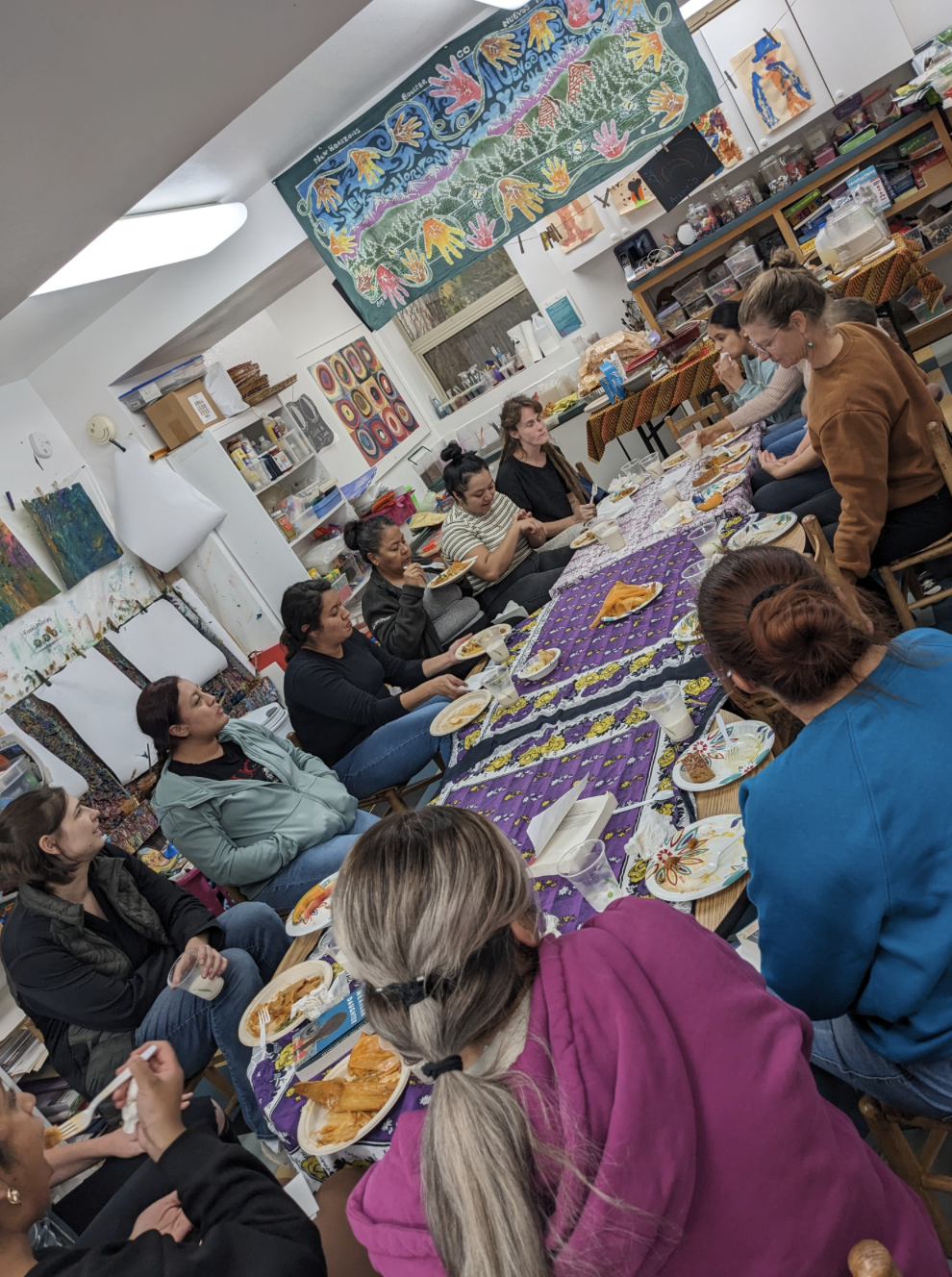 A group of women gathered around a long table enjoying a meal in an art studio or classroom. The table is covered with a purple tablecloth, and the room has colorful artwork and supplies on the walls and shelves.
