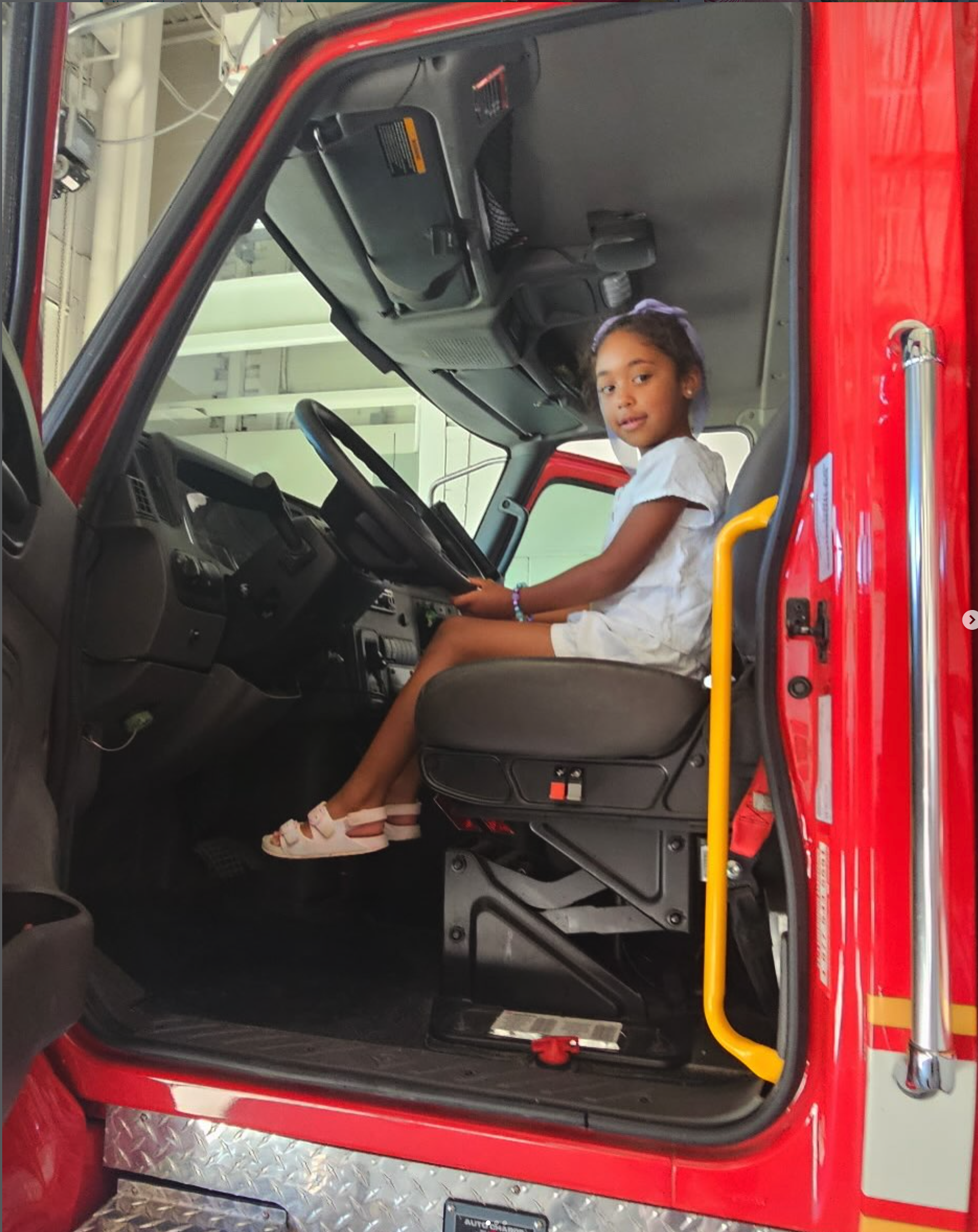 A young girl sitting in the driver's seat of a red fire truck, smiling, with her hands on the steering wheel inside the fire truck's cabin.