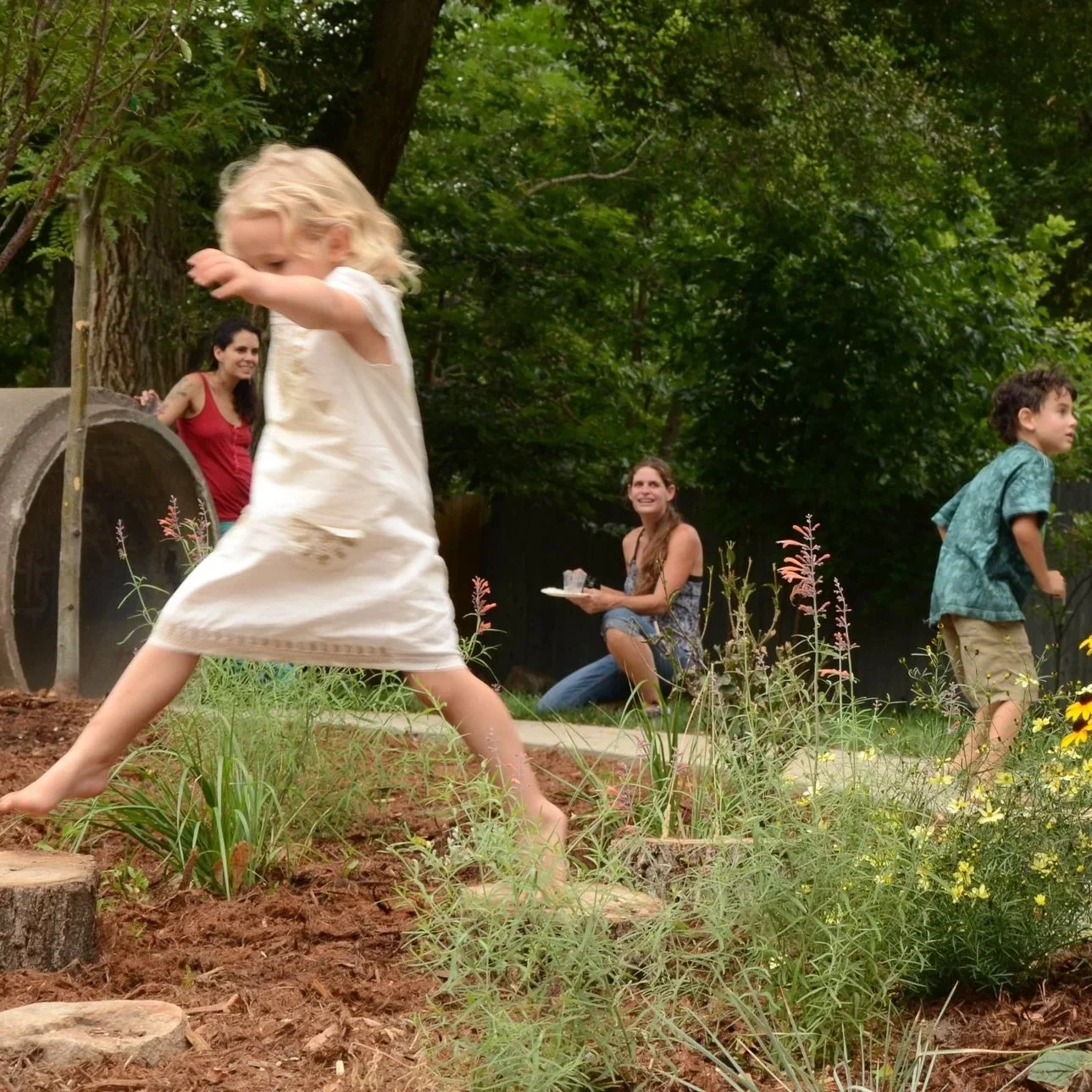 Children playing in the New Horizons play space with two women sitting and smiling in the background, surrounded by trees and plants.