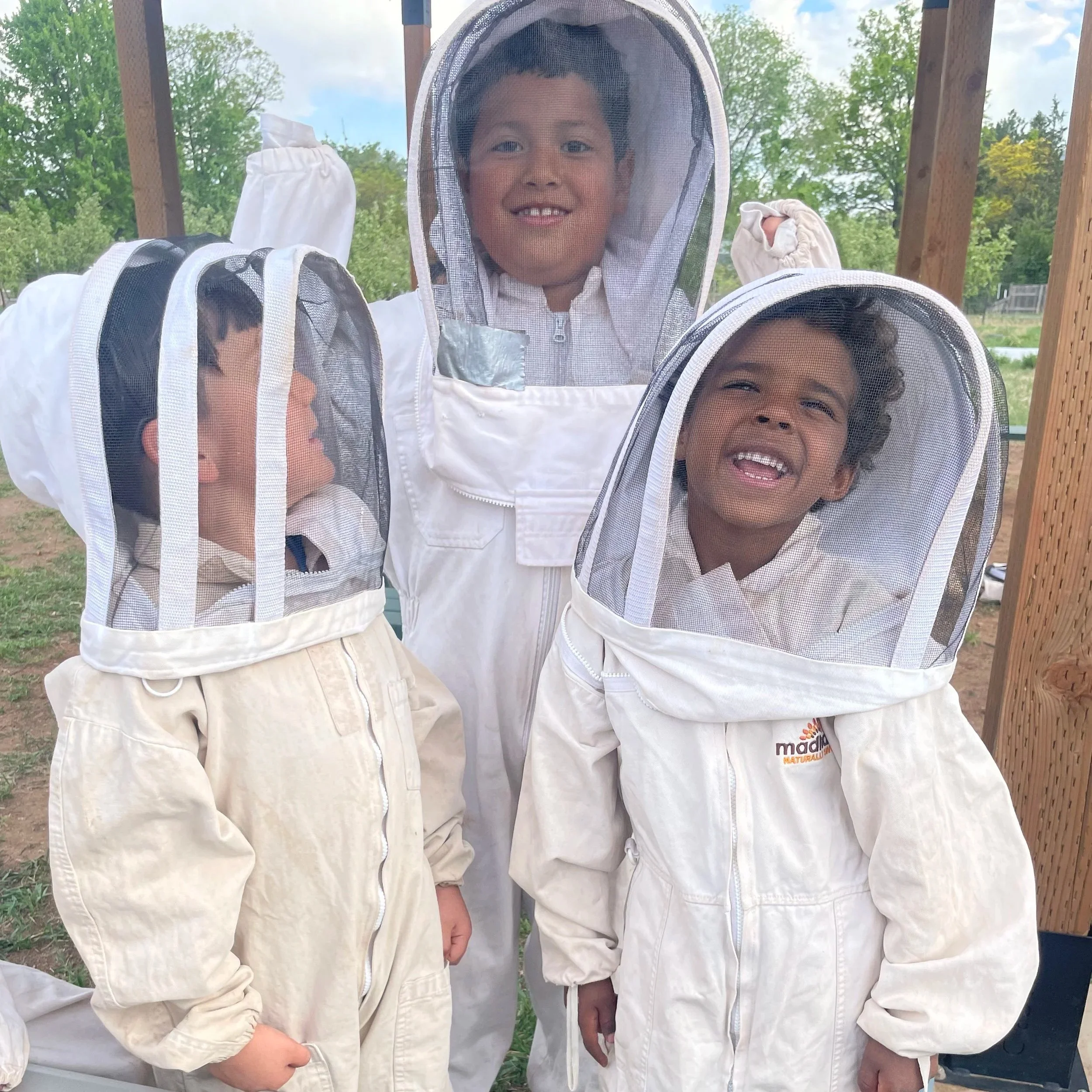 Three children in beekeeping suits and veils standing outdoors, with trees and a wooden structure in the background.
