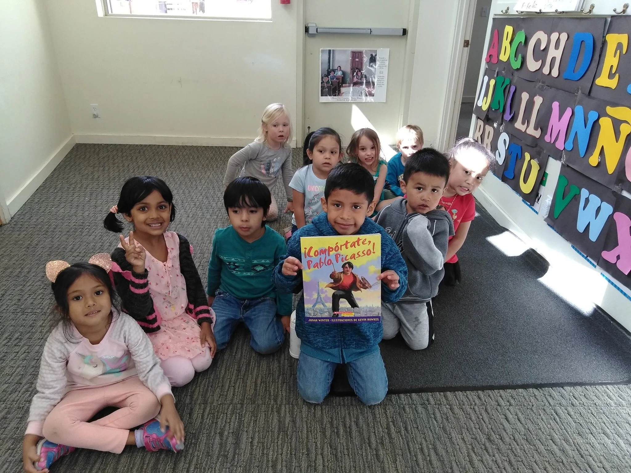 Group of young children sitting on a carpeted floor in a classroom, with colorful letters on the wall, some children smiling, and one child holding a book titled '¡Compórtate, Pablo Picasso!'