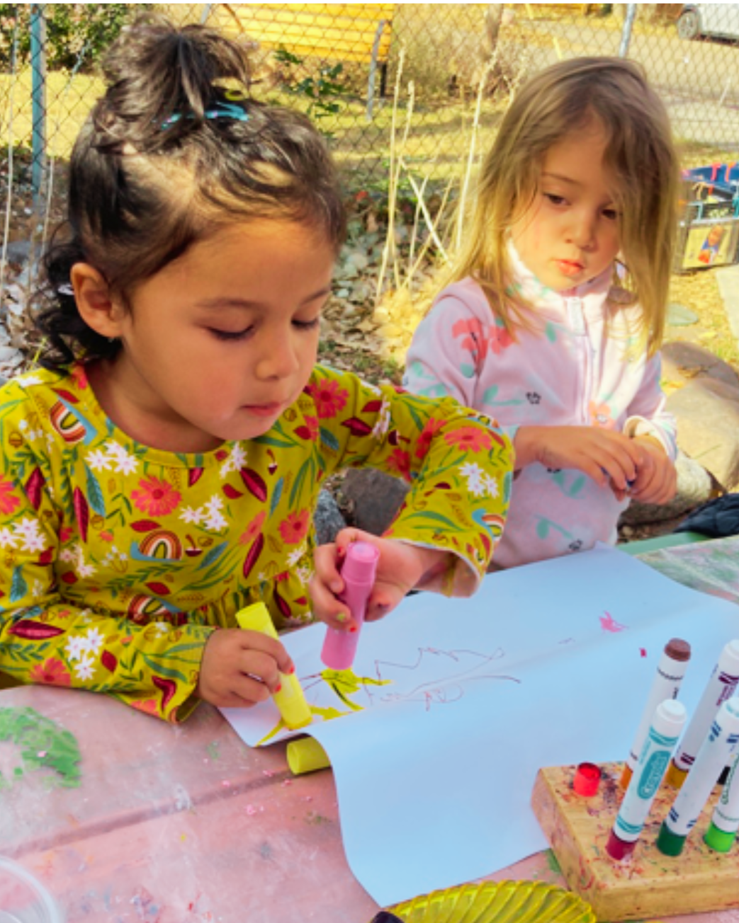 Two young girls are sitting at a table outdoors, drawing with colorful markers on a large sheet of paper. One girl has dark hair styled in a half-up bun with blue and yellow hair accessories, and is wearing a yellow dress with floral and rainbow patterns. The other girl has blonde hair, slightly messy, and is wearing a light pink hoodie with colorful designs. The table is covered with art supplies, and the background includes a chain-link fence and some outdoor greenery.