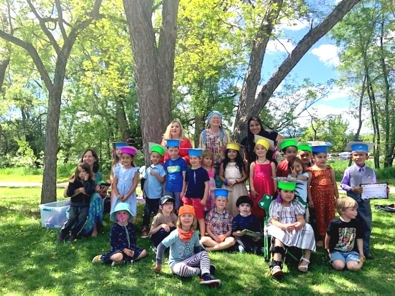 A large group of children and adults outdoors under a large tree on a sunny day, some children wearing colorful preschool graduation hats, in a park-like setting.
