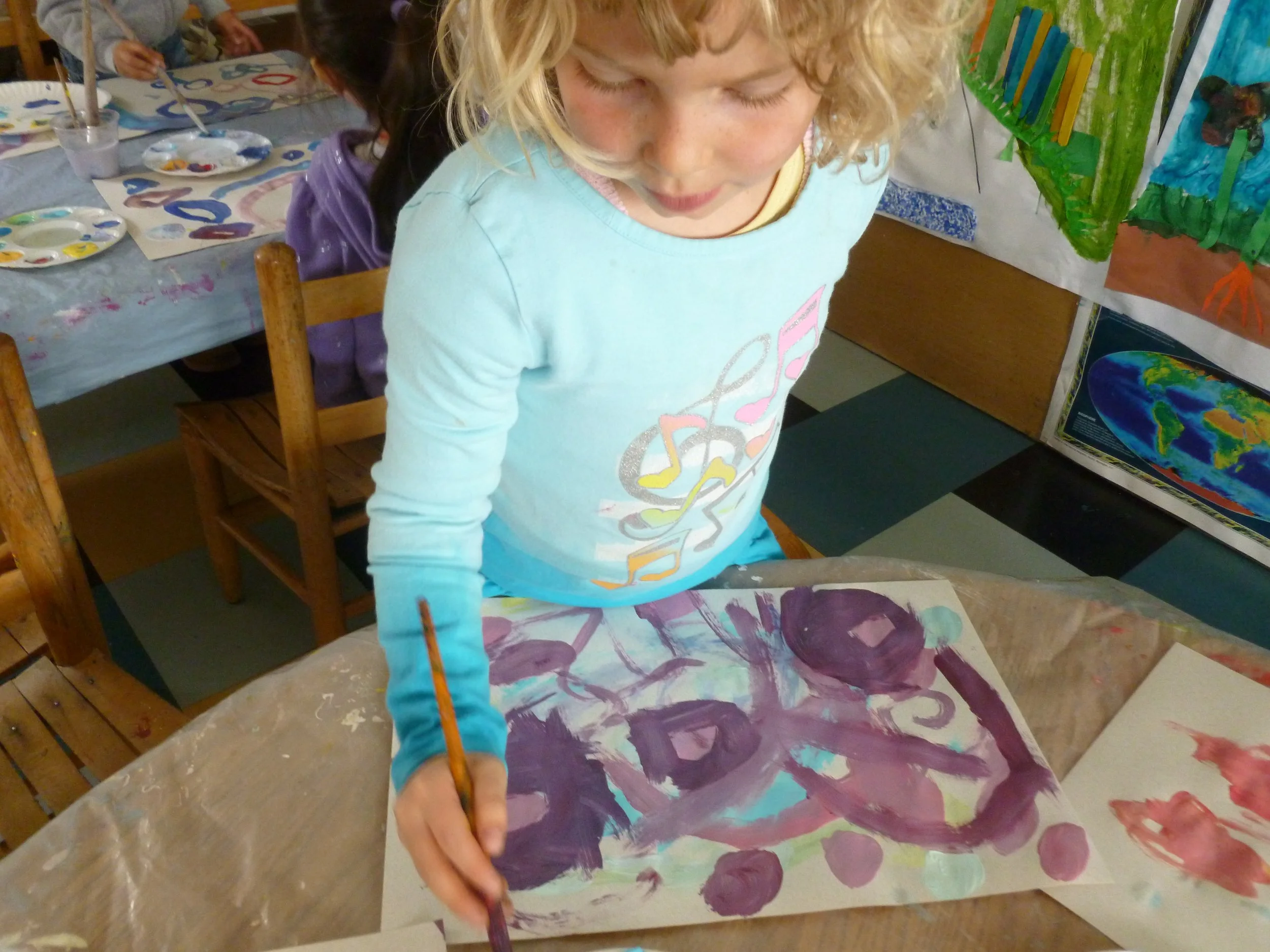 A young girl with curly blonde hair painting on a large sheet of paper with purple, pink, and teal colors in an art classroom.